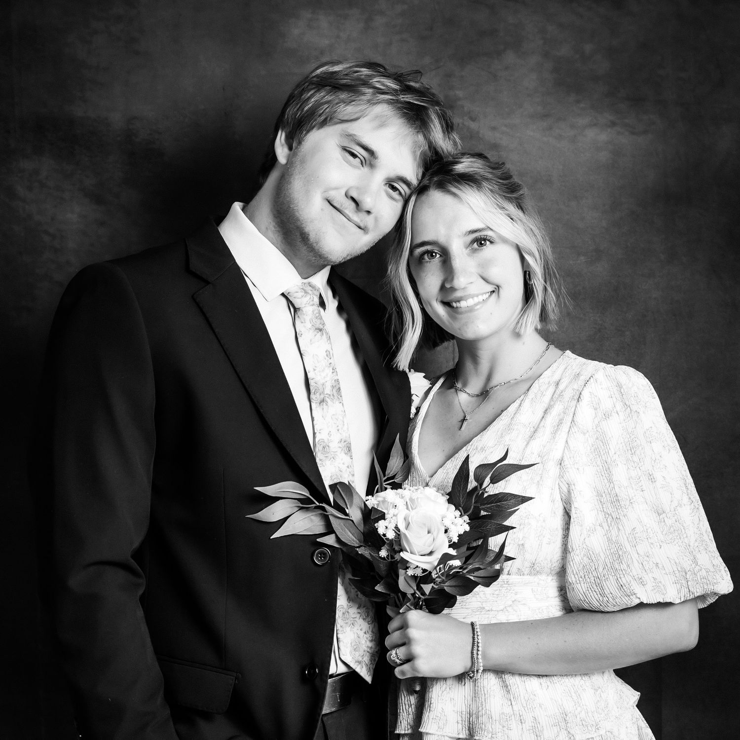 Black and white portrait of a smiling couple, man in suit and woman in white dress holding a floral bouquet.