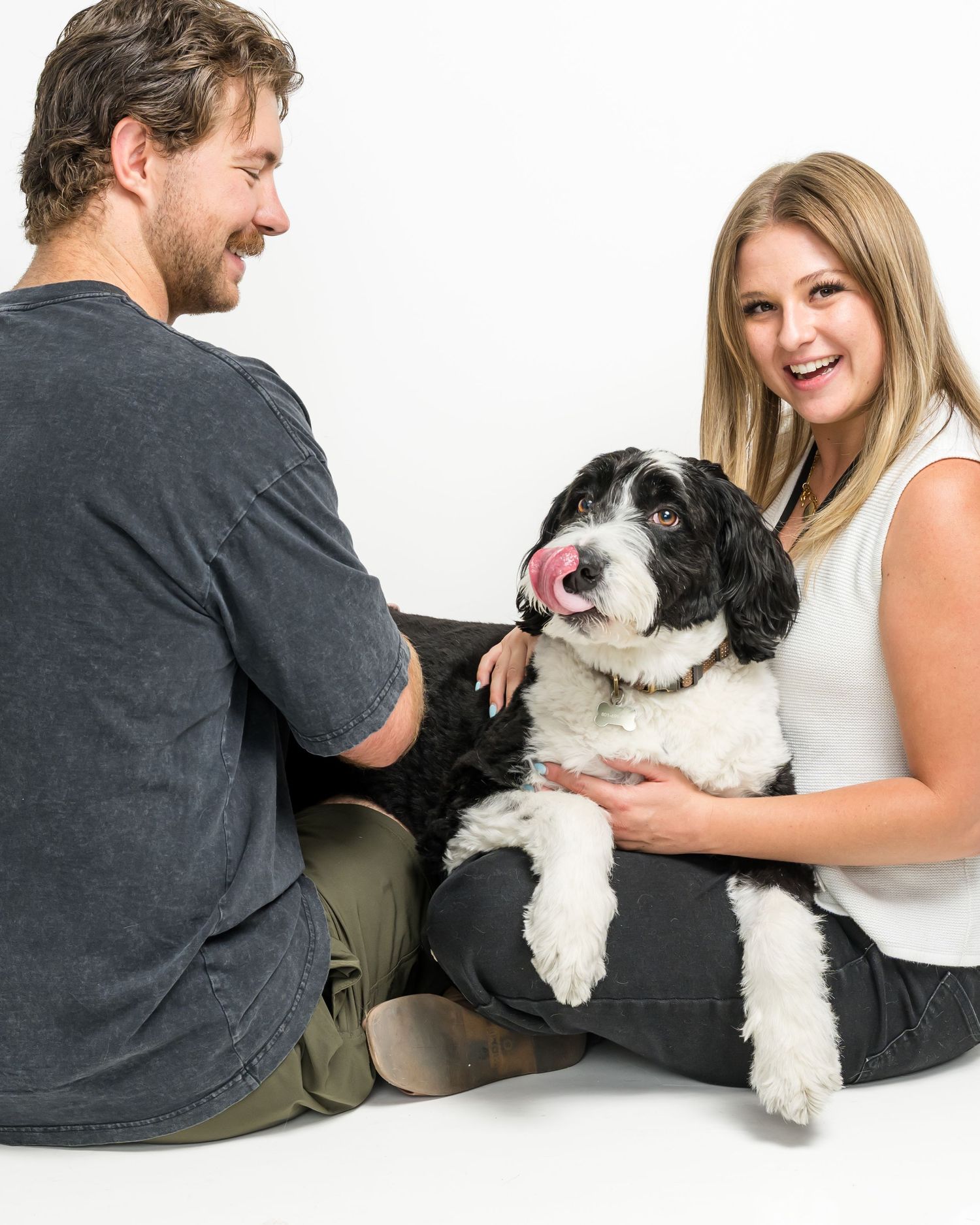 Couple sitting on white background with black and white Cockapoo dog licking its nose while being petted.