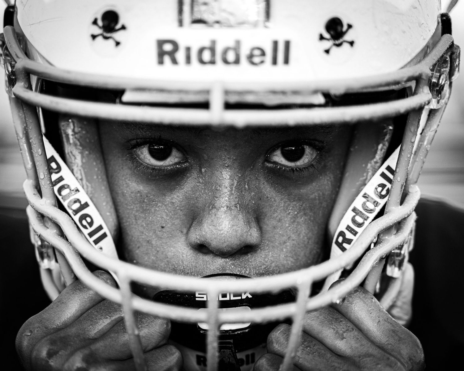 Close-up black and white photo of a young football player wearing a Riddell helmet with intense focused eyes.