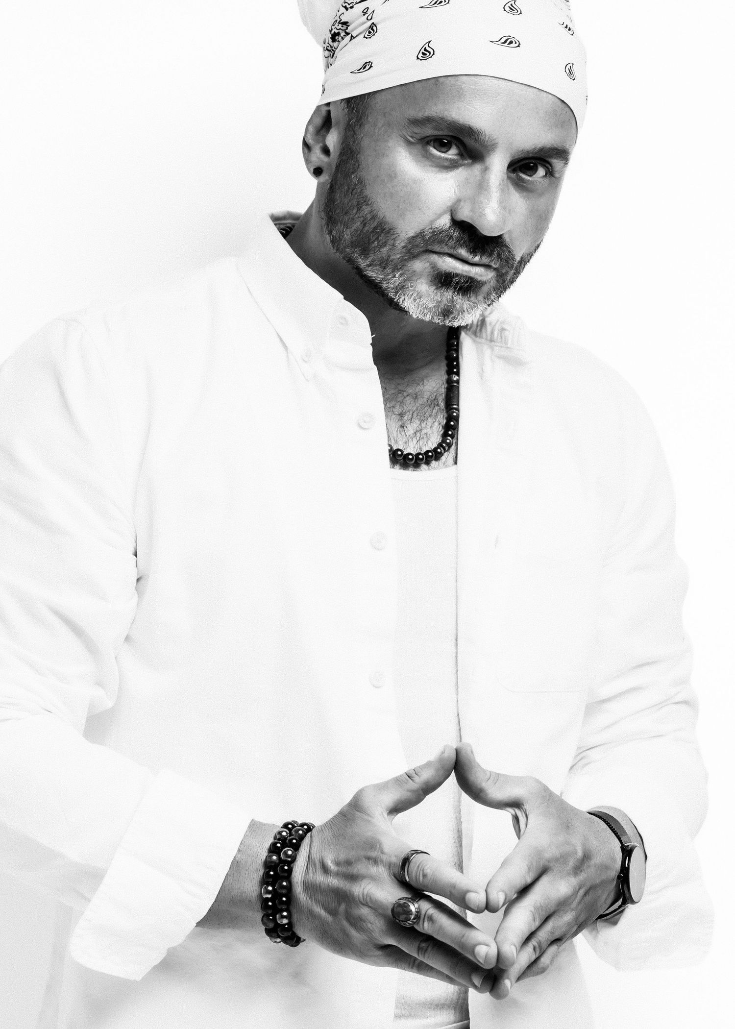 Black and white portrait of a bearded man in white shirt and bandana, hands clasped, wearing beaded jewelry.
