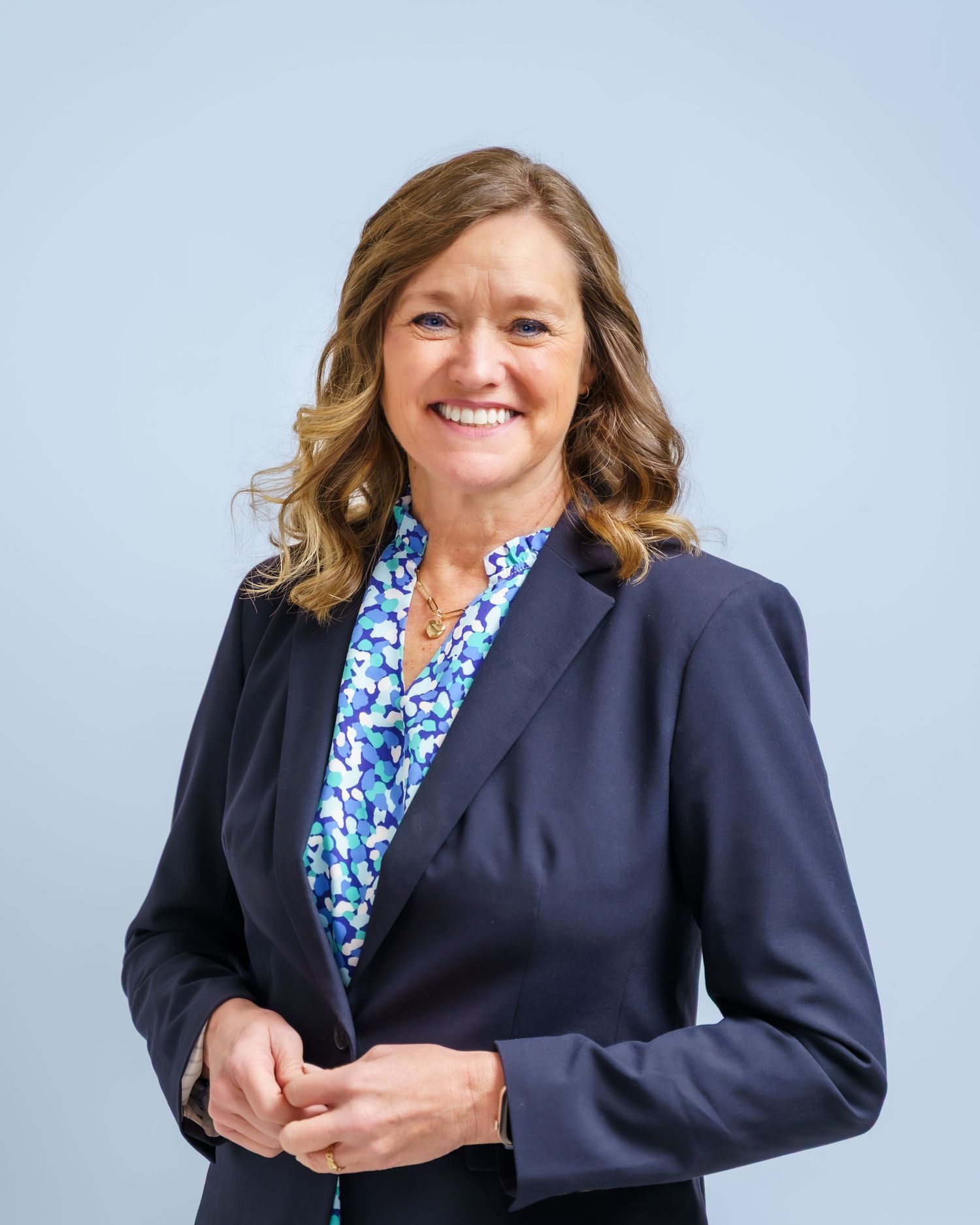 Professional woman in navy blazer and floral blouse smiling confidently against a light blue background.