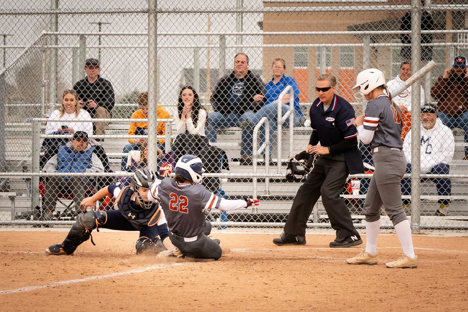Softball player sliding into home plate as catcher attempts tag while umpire watches during competitive game.