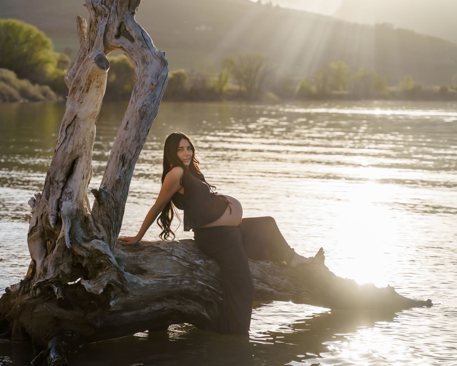 Woman in black outfit posing on driftwood by a sunlit river with green hills in the background.