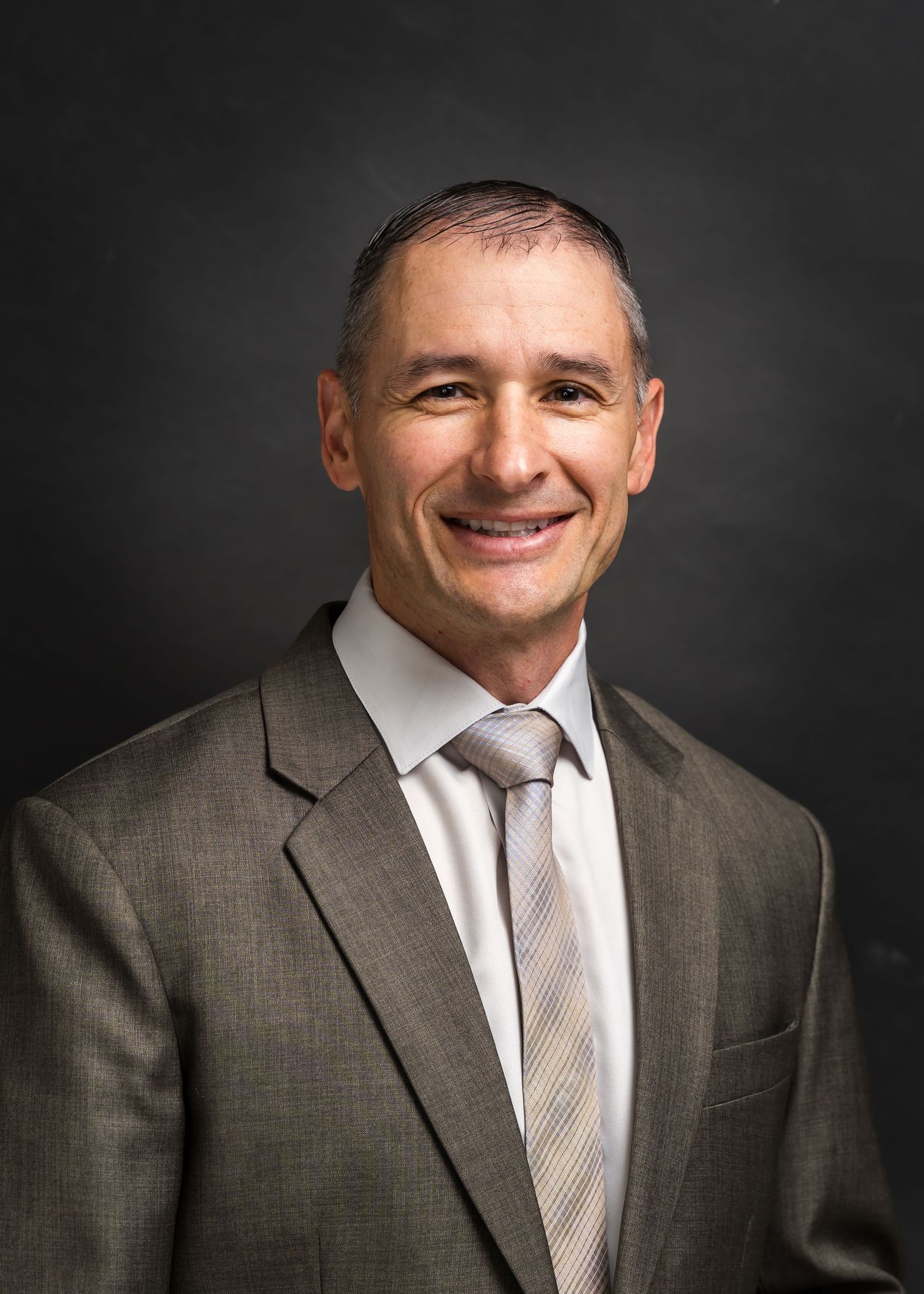 Professional headshot of a smiling middle-aged man in a gray suit and striped tie against a dark background.