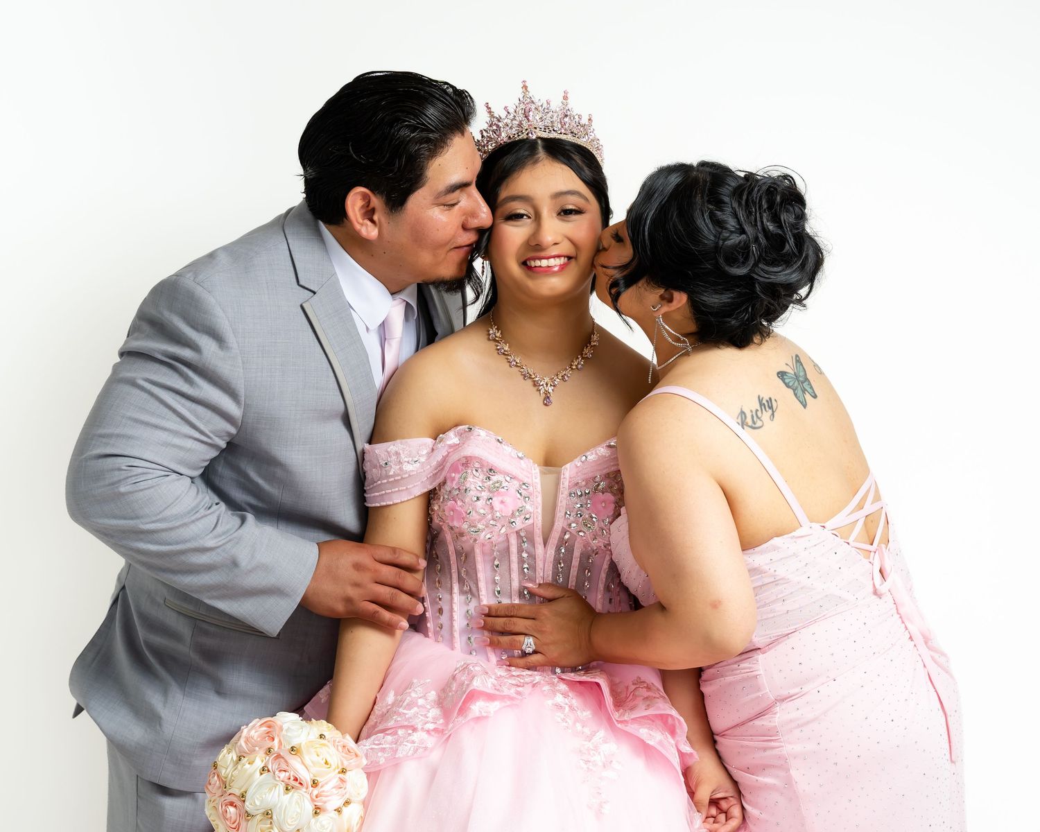 A young woman in a pink quinceañera gown with tiara receives kisses from her parents in a studio portrait.