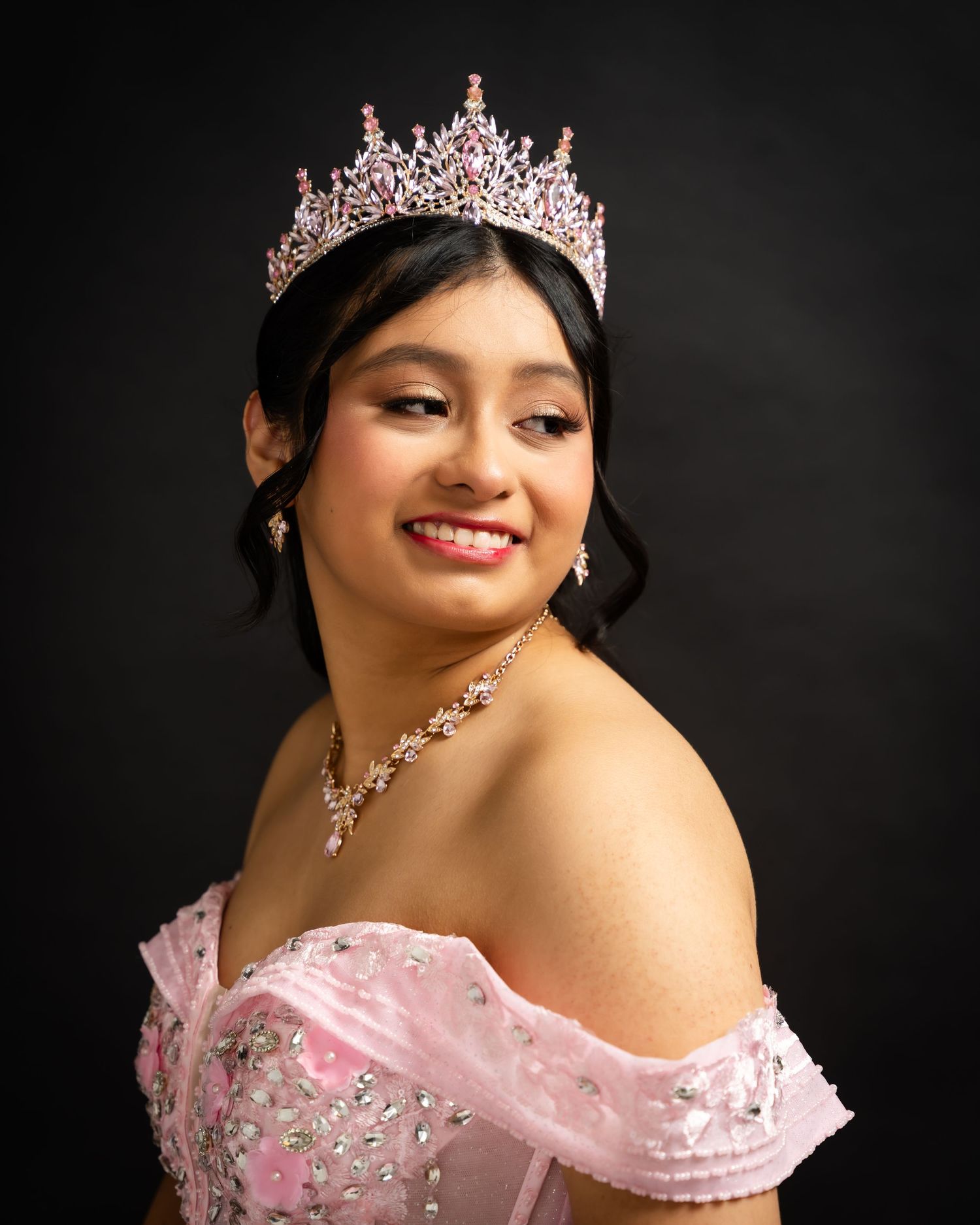 Young woman in pink off-shoulder quinceañera gown wearing a sparkling tiara and jewelry, smiling against dark background.