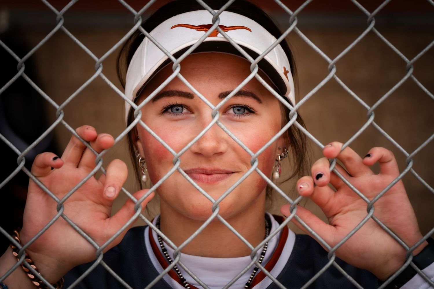 Female softball player in uniform grips chain-link fence, looking through it with blue eyes and a white visor.