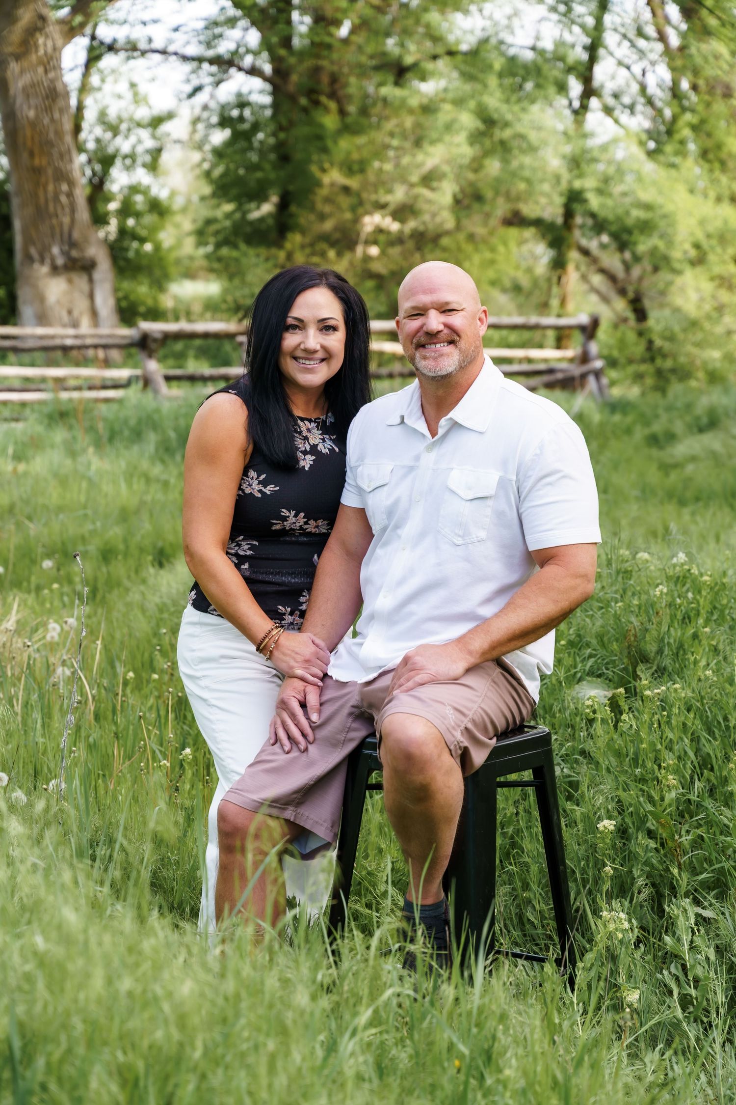 A smiling couple poses together in a lush green meadow, the woman in a floral top and the man in a white polo shirt.