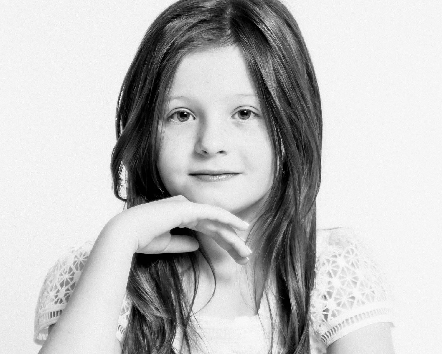 Young girl with long brown hair poses thoughtfully with chin on hand in black and white portrait.