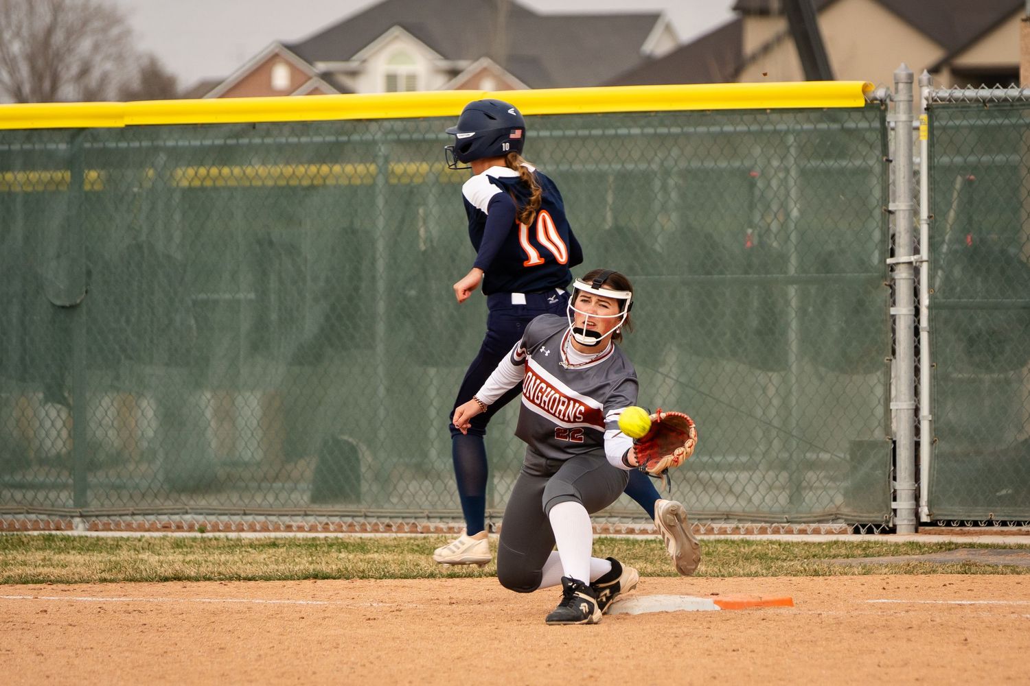 Softball player catches ball at base during youth league game on dirt field with chain-link fence background.