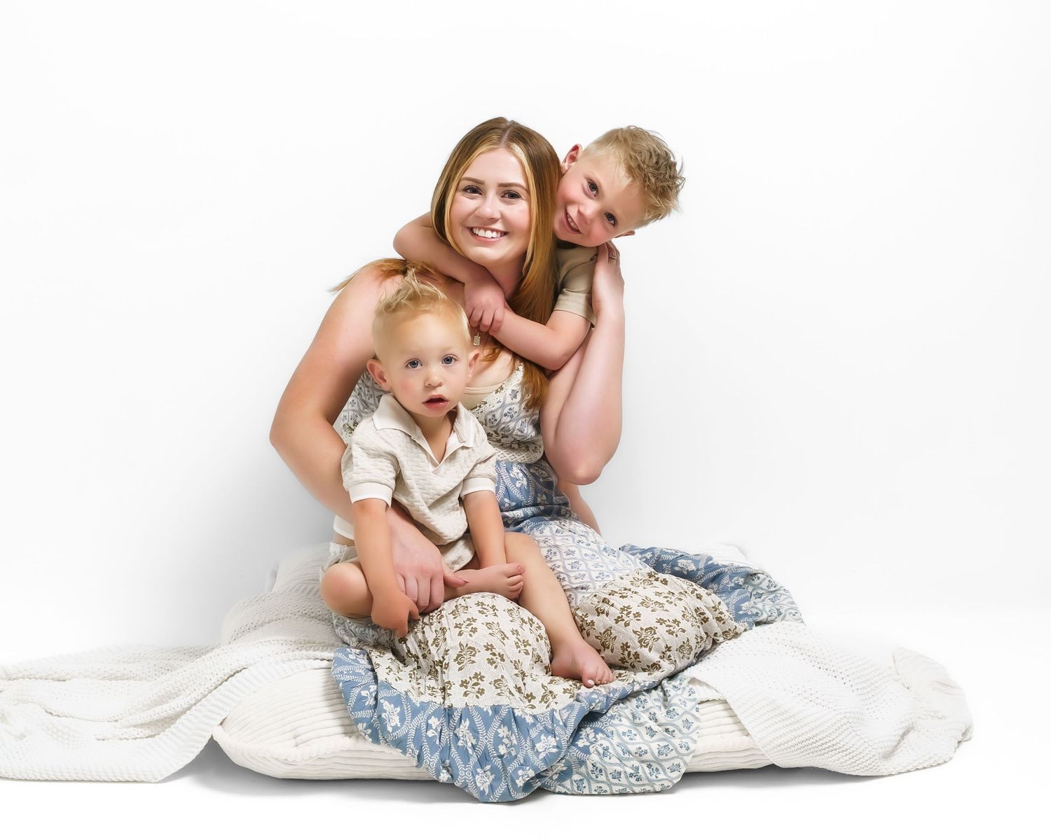 Joyful mother in floral dress hugging two young sons on white blankets in studio portrait.