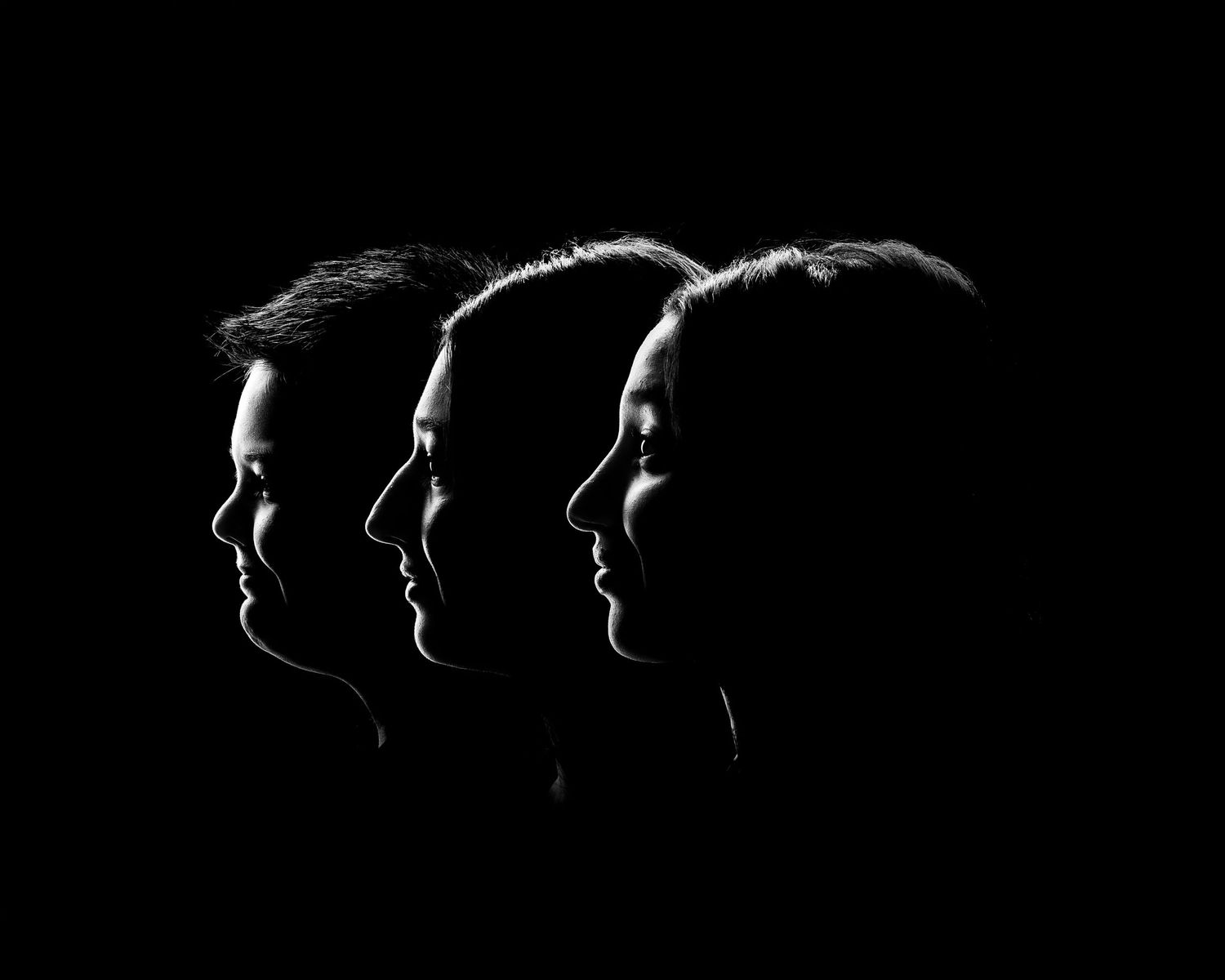 Three people in silhouette profile against black background, dramatic low-key black and white portrait photography.