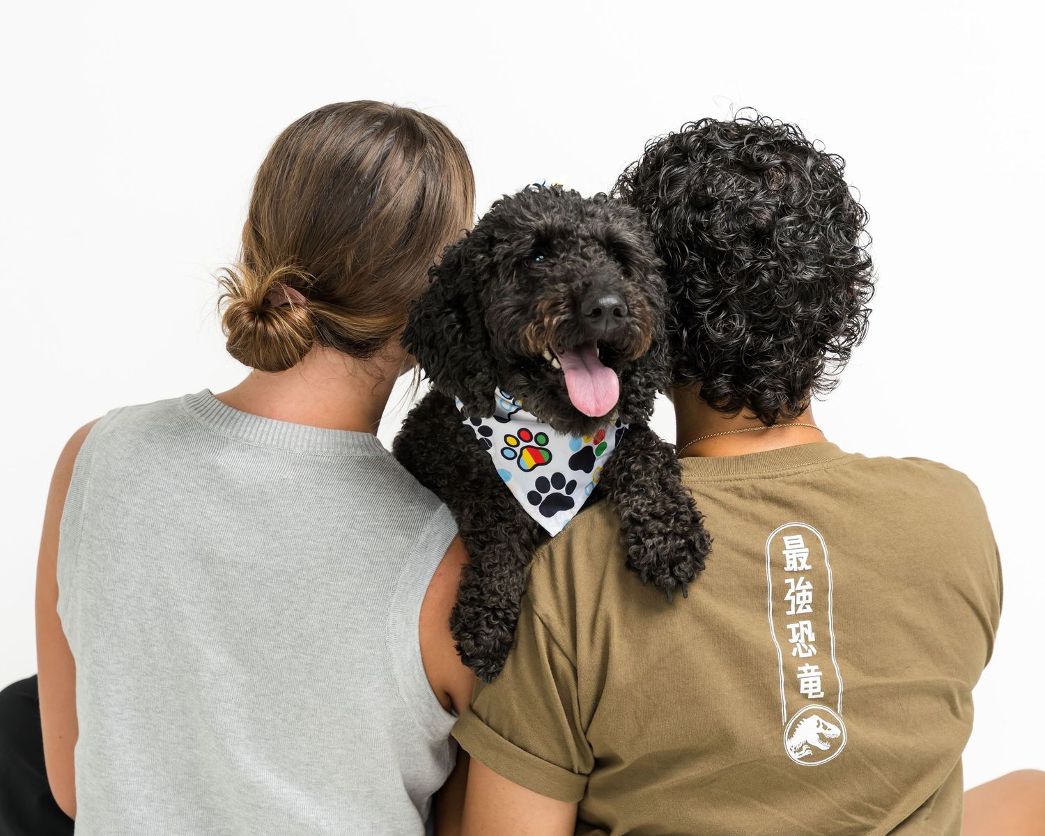 Two people sitting with a black curly-haired dog wearing a colorful paw print bandana between them.