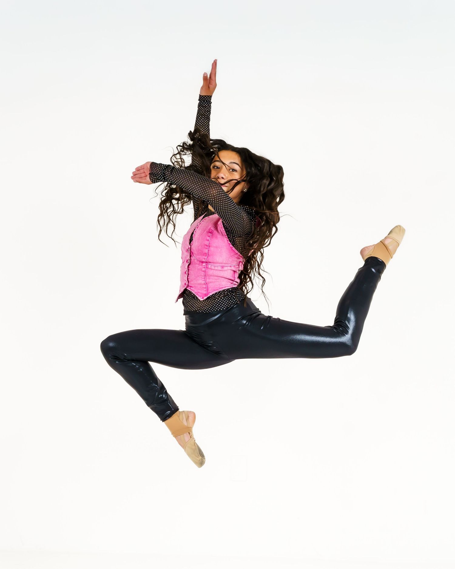A dancer leaps mid-air in a pink top and shiny black leggings, arms raised, long curly hair flowing, against a white background.