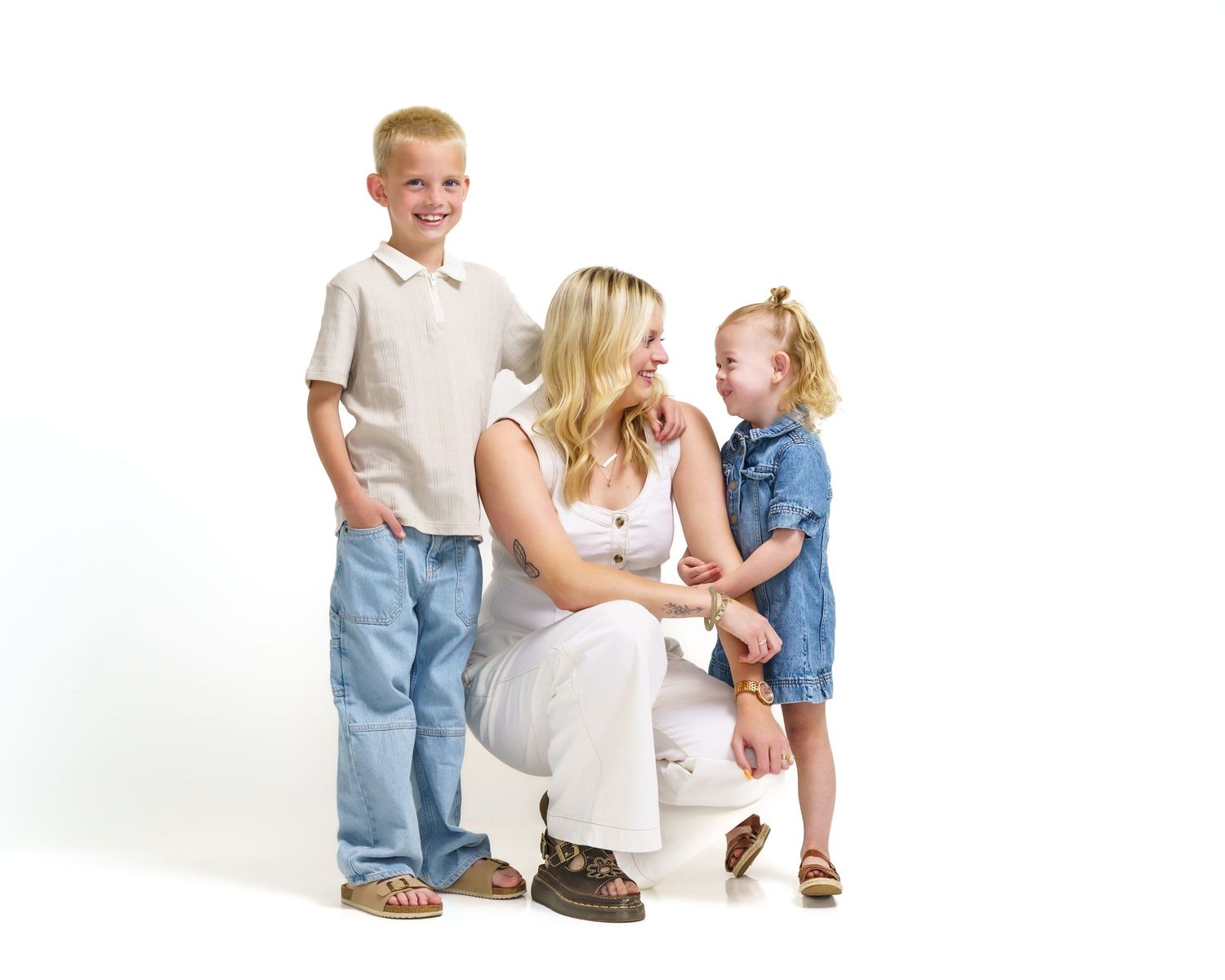 Mother crouching between laughing son and toddler daughter in bright white studio portrait.