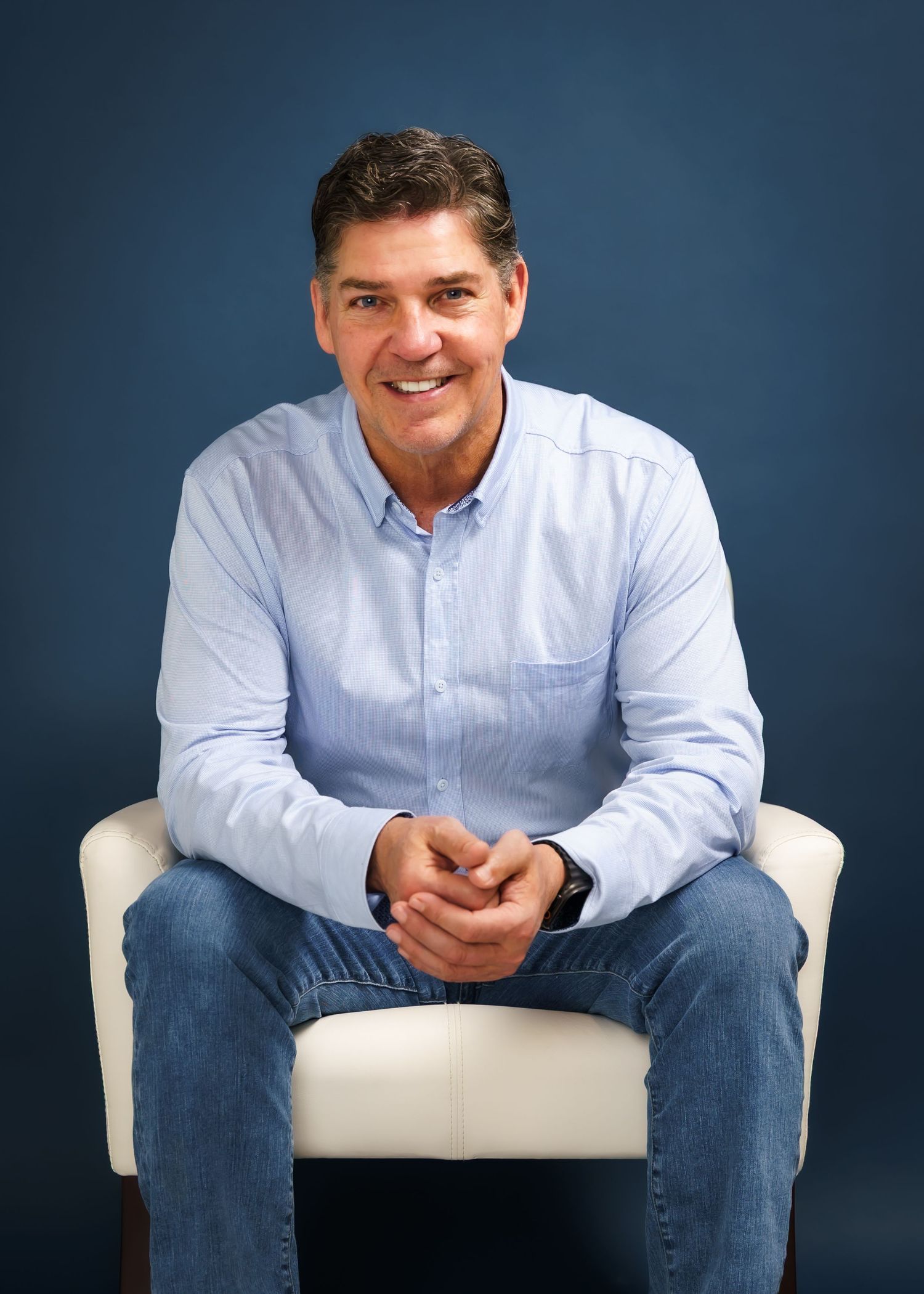Middle-aged man in light blue shirt and jeans sitting relaxed on white chair, smiling against dark blue background.