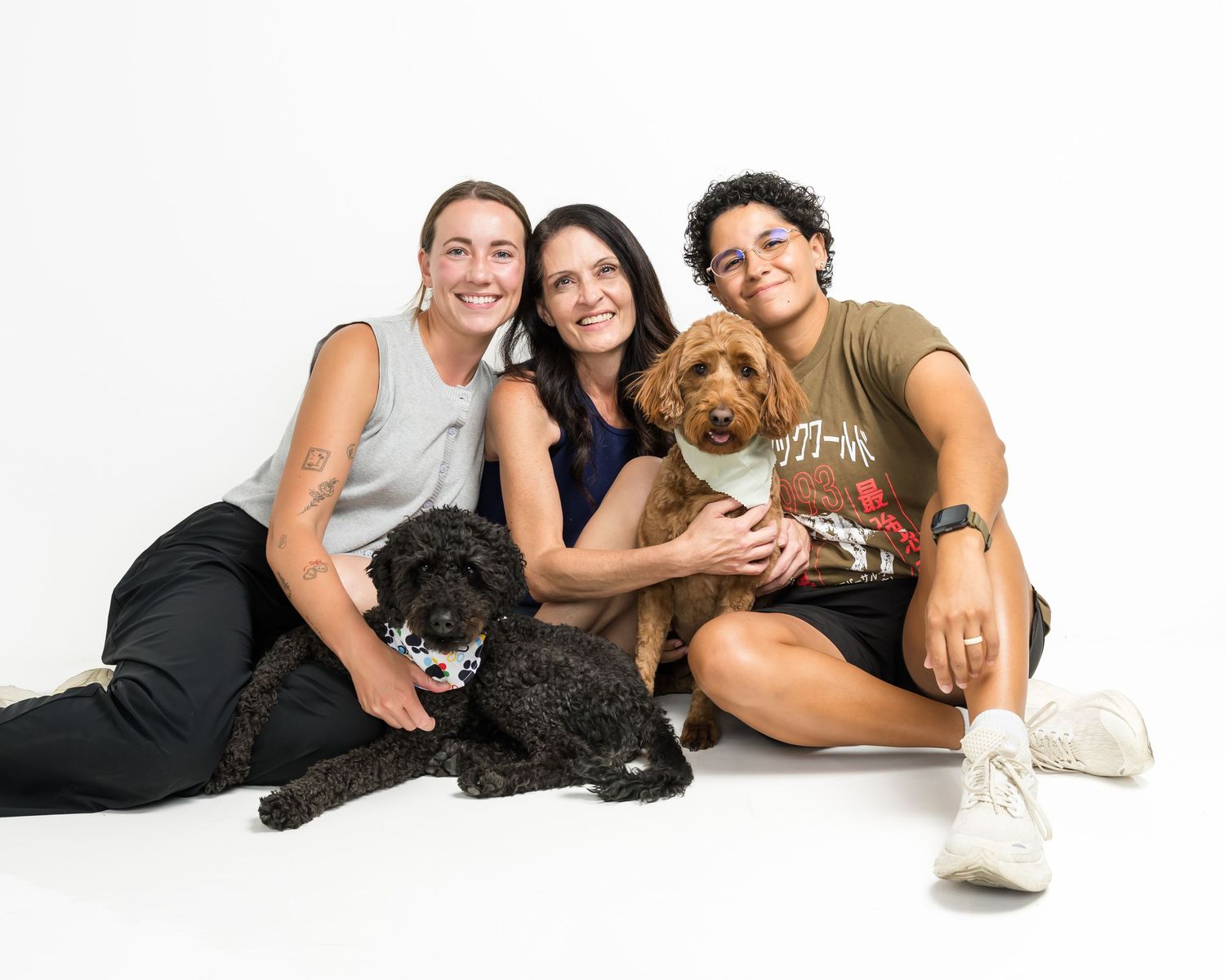 Three women smile together on white background with two dogs, a black poodle and a golden doodle, in a studio portrait.