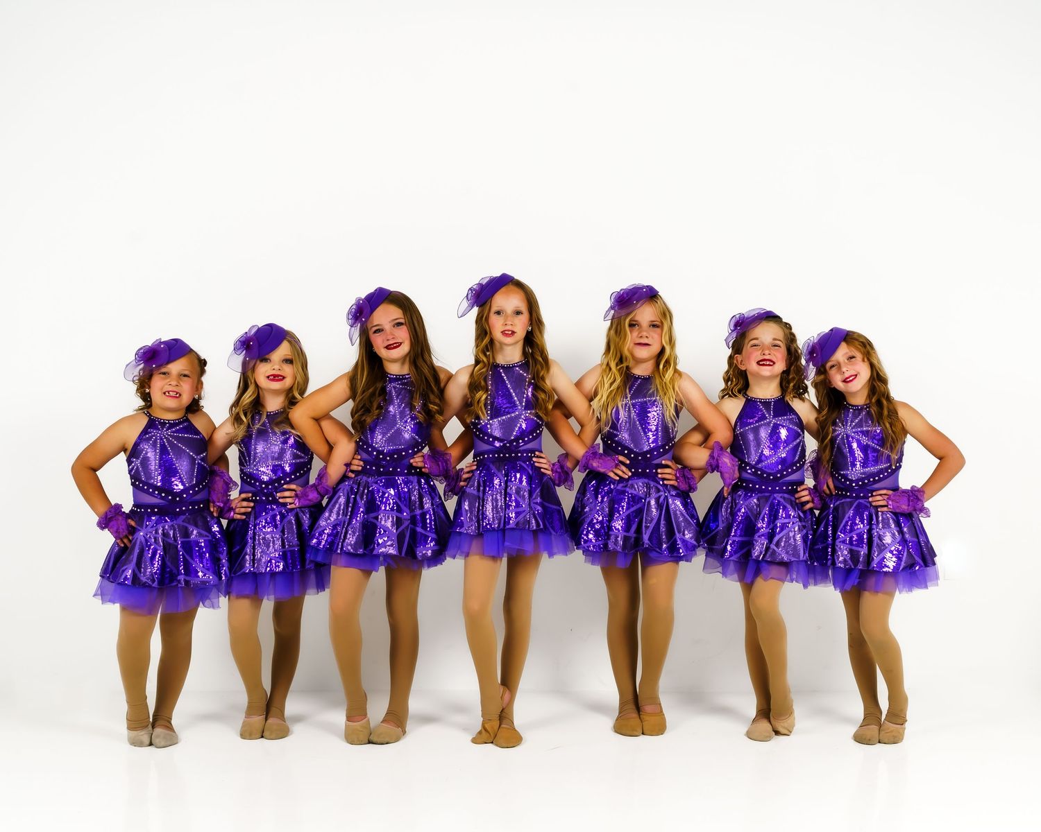 Seven young girls in purple sequin dance costumes and hats posing together for a dance recital photo.