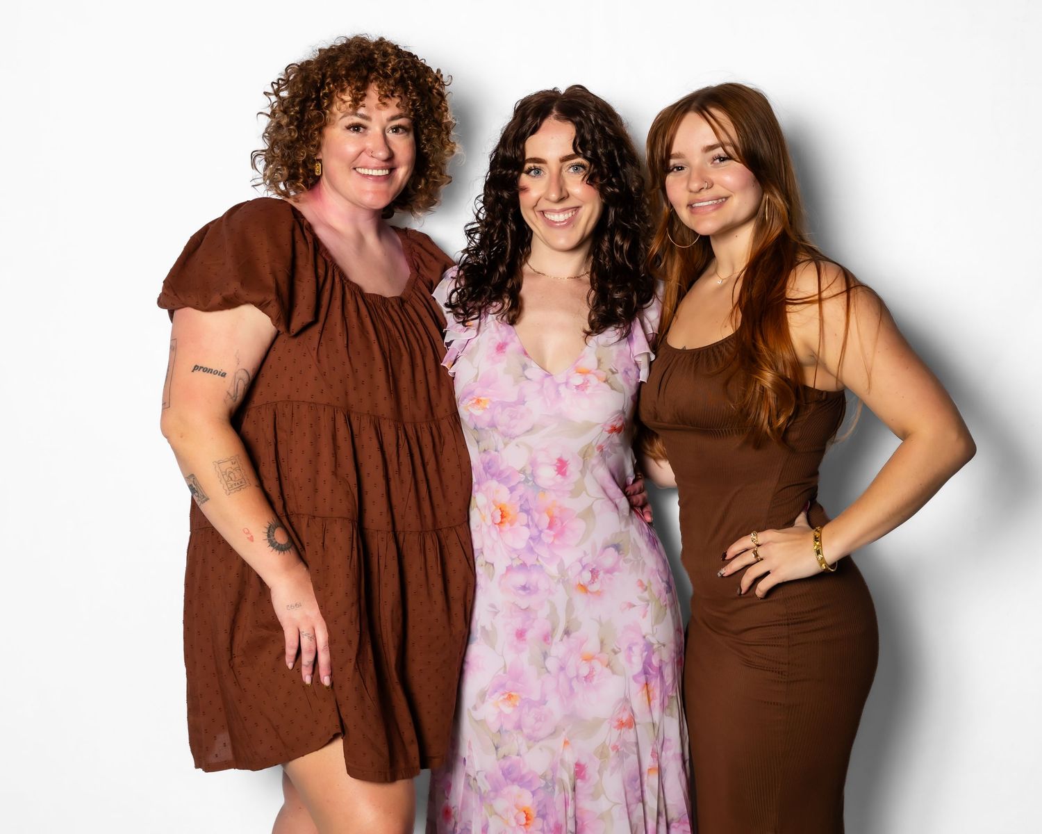 Three women smiling together in brown and floral outfits against a white background.