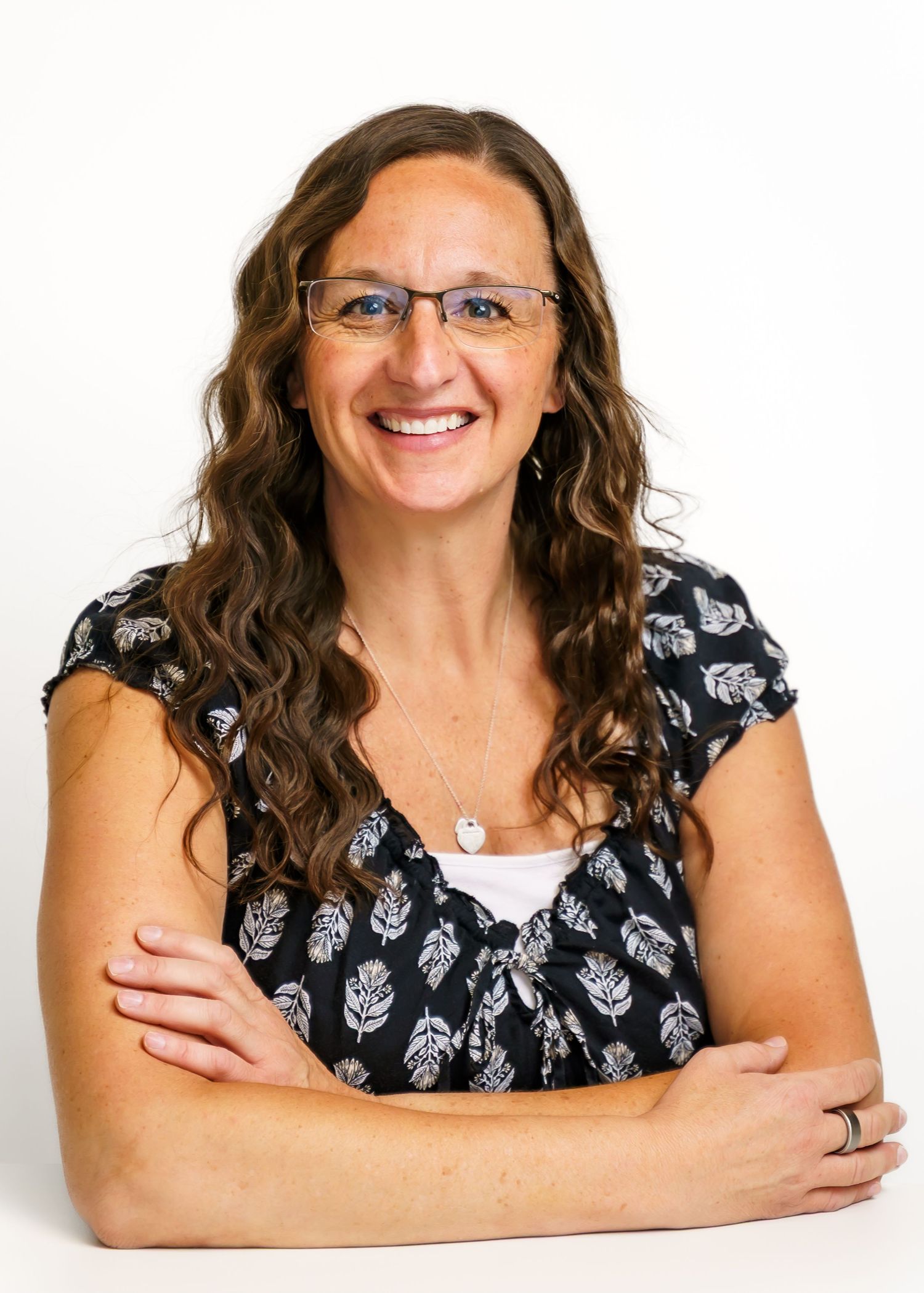Smiling woman with curly brown hair and glasses, arms crossed, wearing a floral top in a professional headshot.