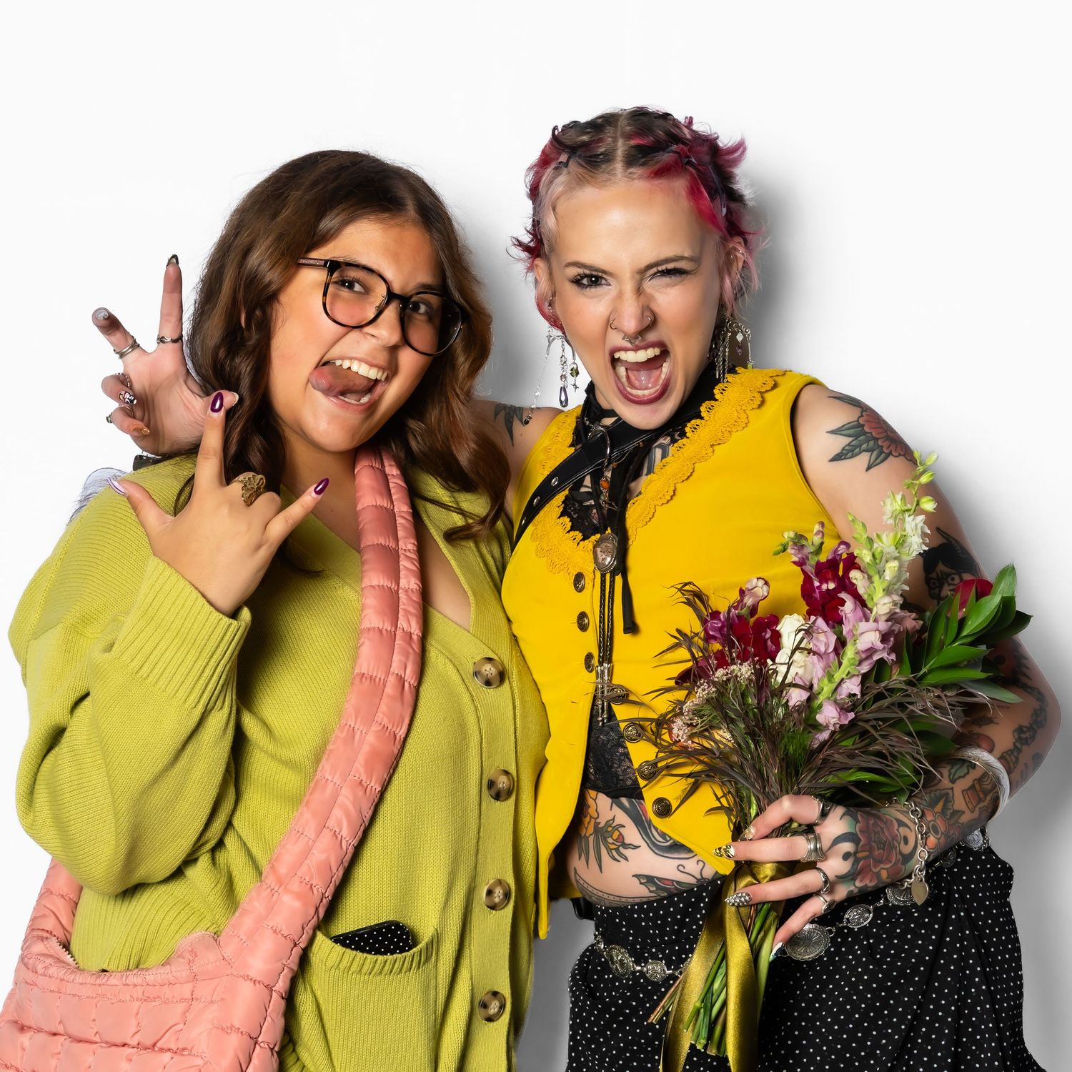 Two tattooed women pose together, one in green cardigan with glasses, other in yellow vest holding flowers, against white background.