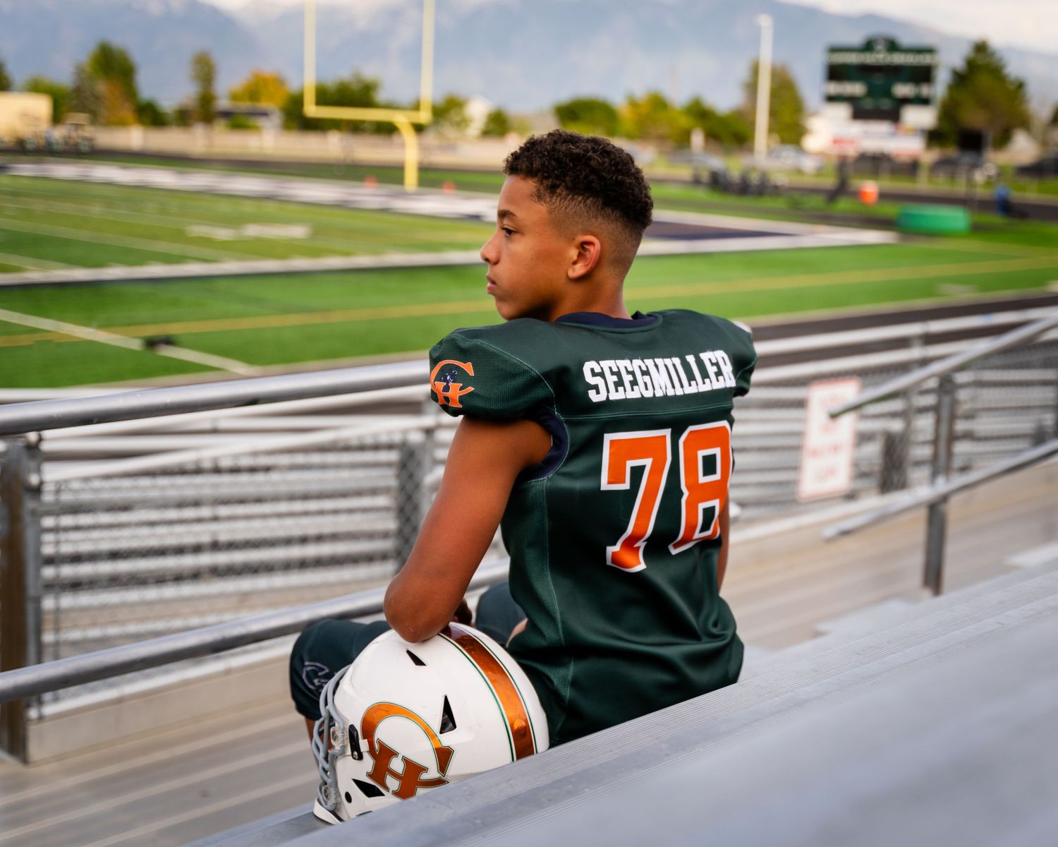 Young football player in green Seemiller #78 jersey holds helmet in stadium bleachers with mountains in background.