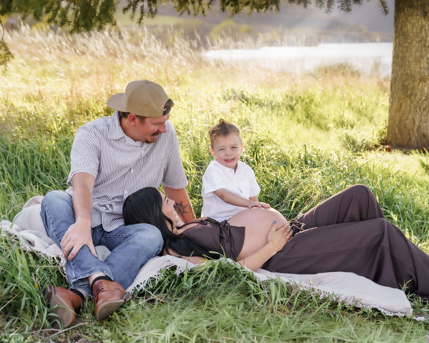 Family maternity photo session outdoors on blanket in green grass, father and toddler smiling at pregnant mother.
