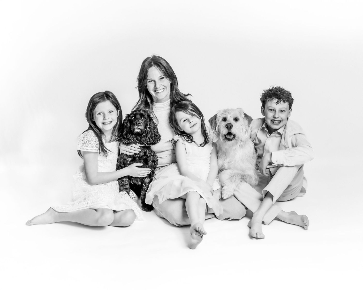 Black and white family portrait with mother, three children, and two dogs posing together on white background.