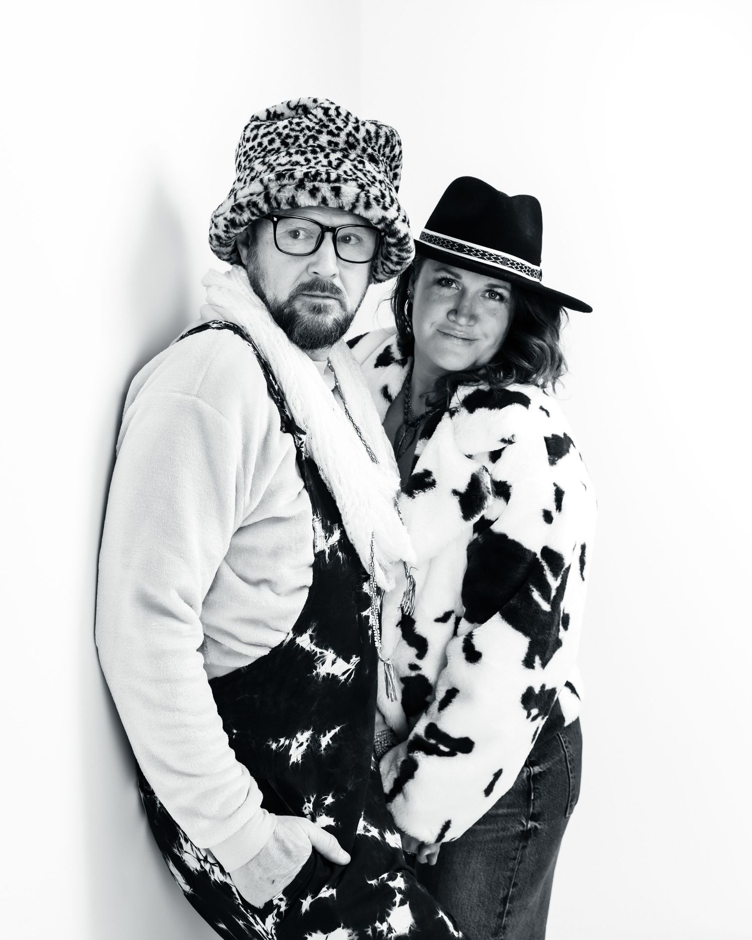 Black and white photo of two people in fun hats and cow print fur coats posing together against a white wall.