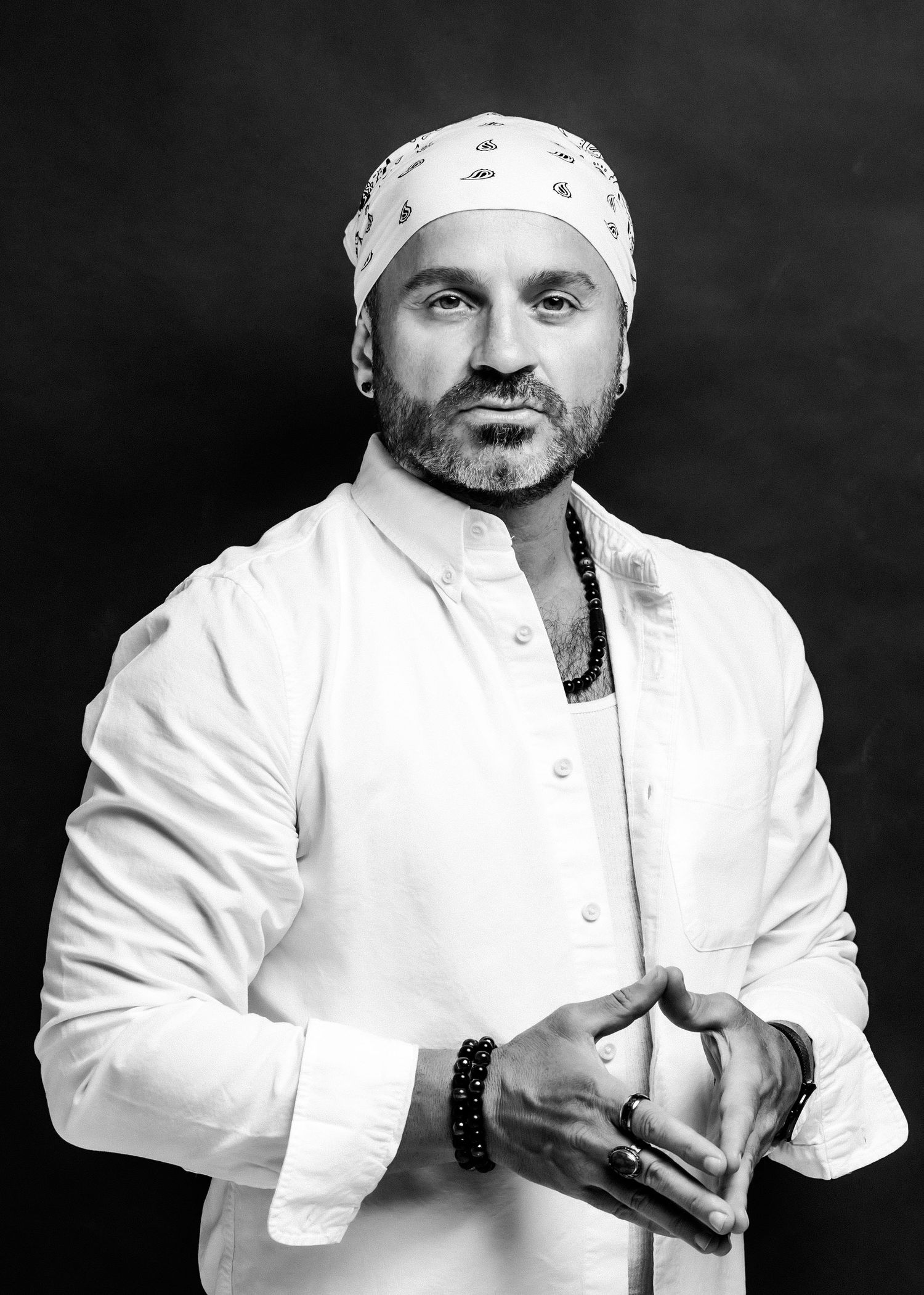 Black and white portrait of a man in white shirt and head wrap, clasping hands, wearing beaded jewelry.