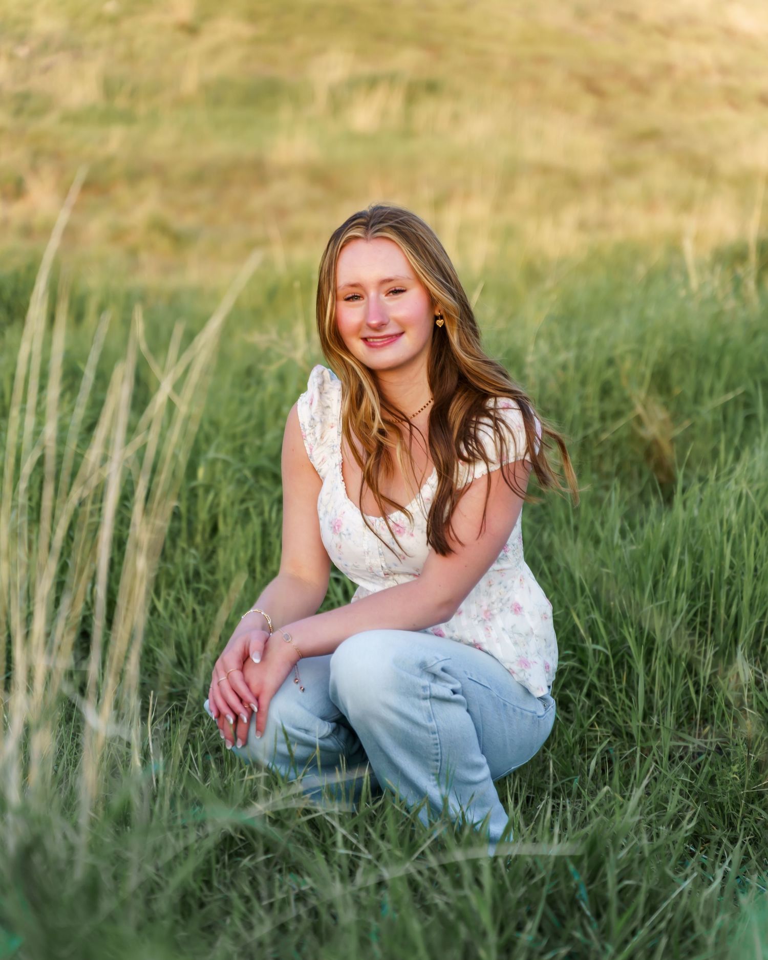 Young woman in floral top and jeans crouching in lush green grass field during golden hour.