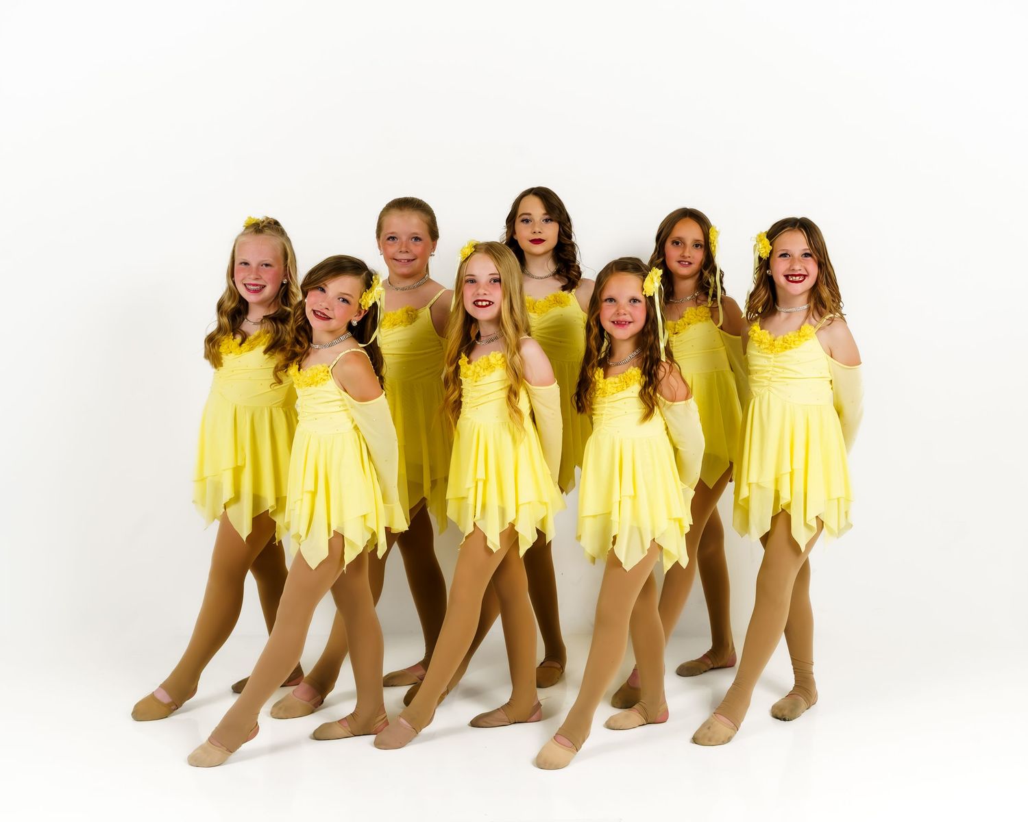 Eight young dancers in matching yellow dresses and nude tights pose together smiling against a white background.