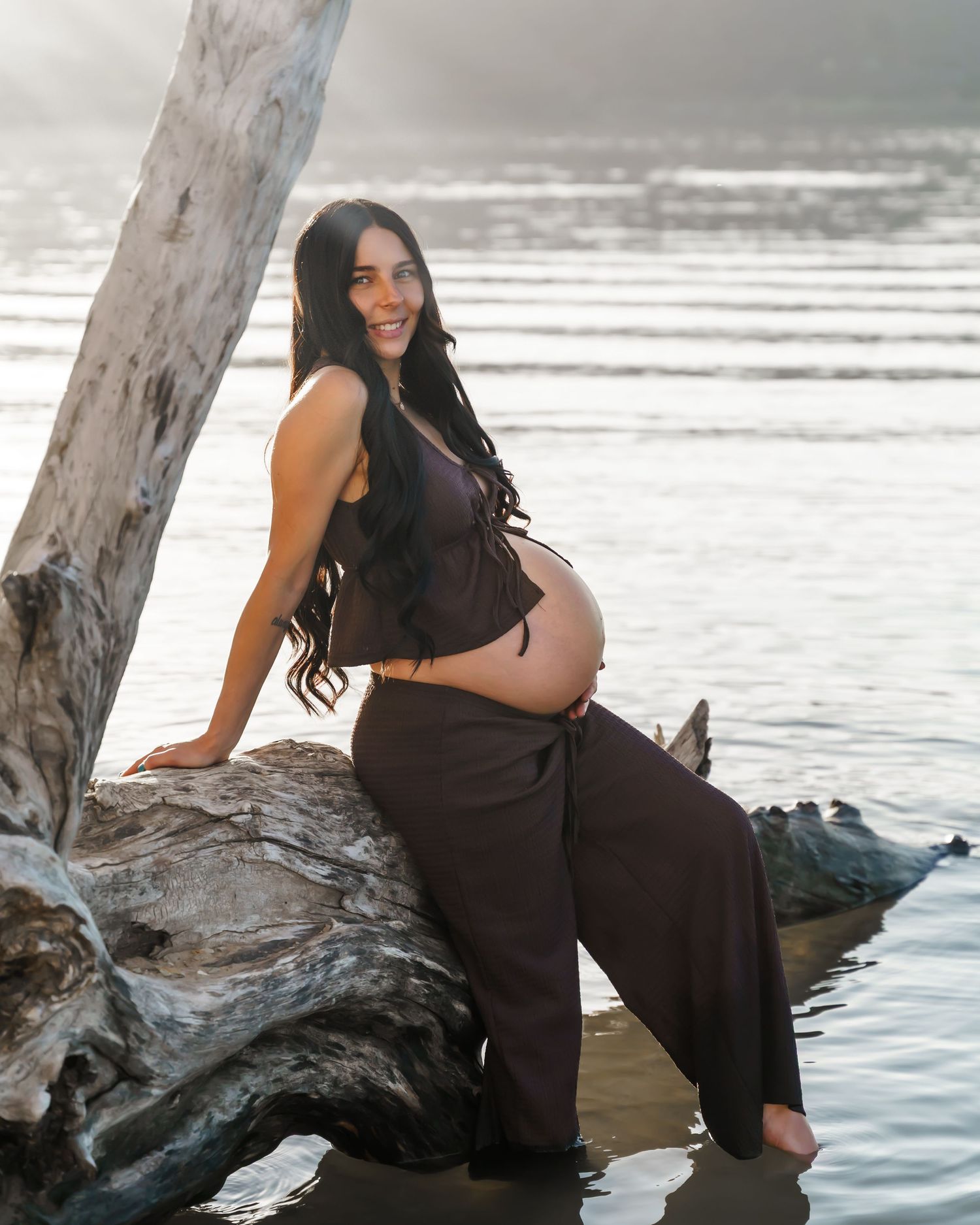 Pregnant woman in dark outfit sitting on driftwood by the water, smiling at camera during golden hour maternity photoshoot.