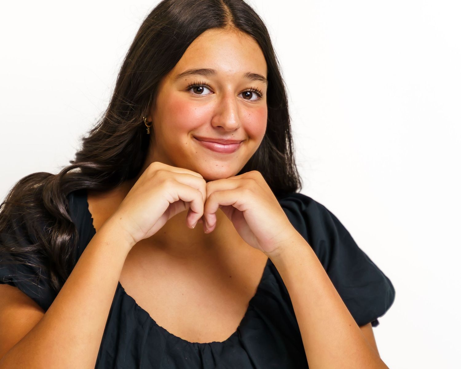 Smiling teenage girl with long dark hair posing with hands under chin against white background.
