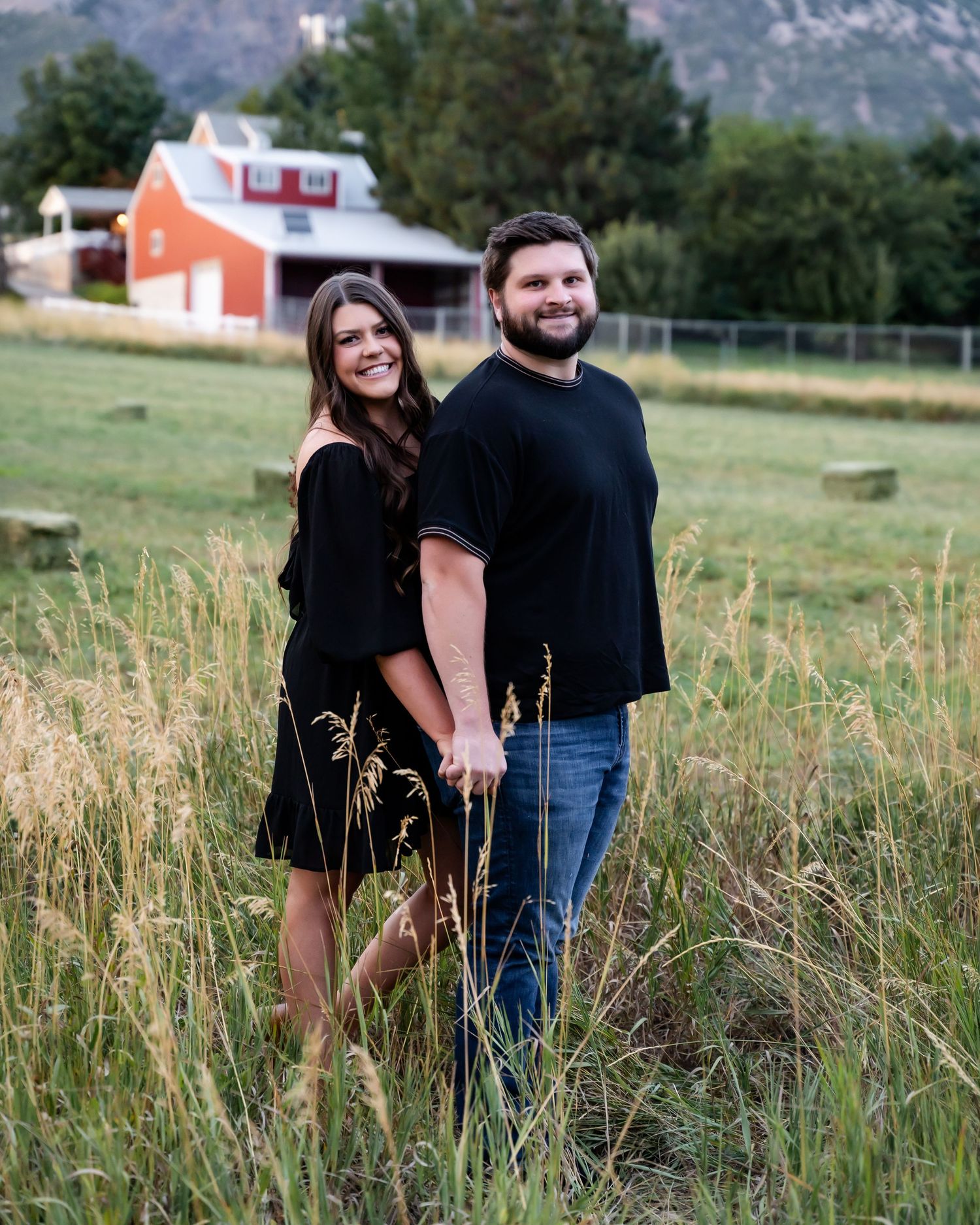 Couple holding hands smiling in a green field with a red barn and mountains in the background.