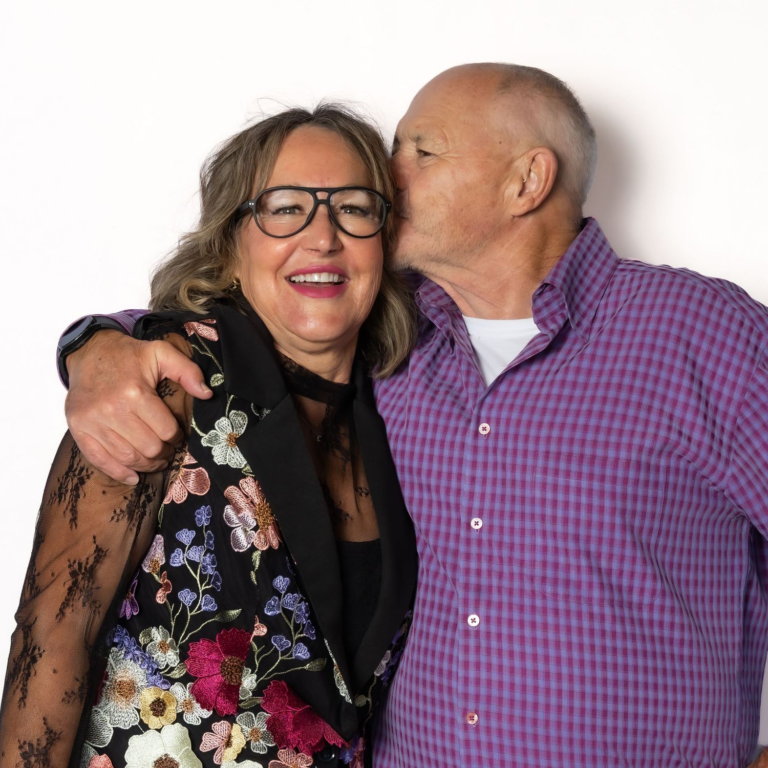 An older couple posing together, the man kissing the woman on the cheek, both smiling warmly against a white background.
