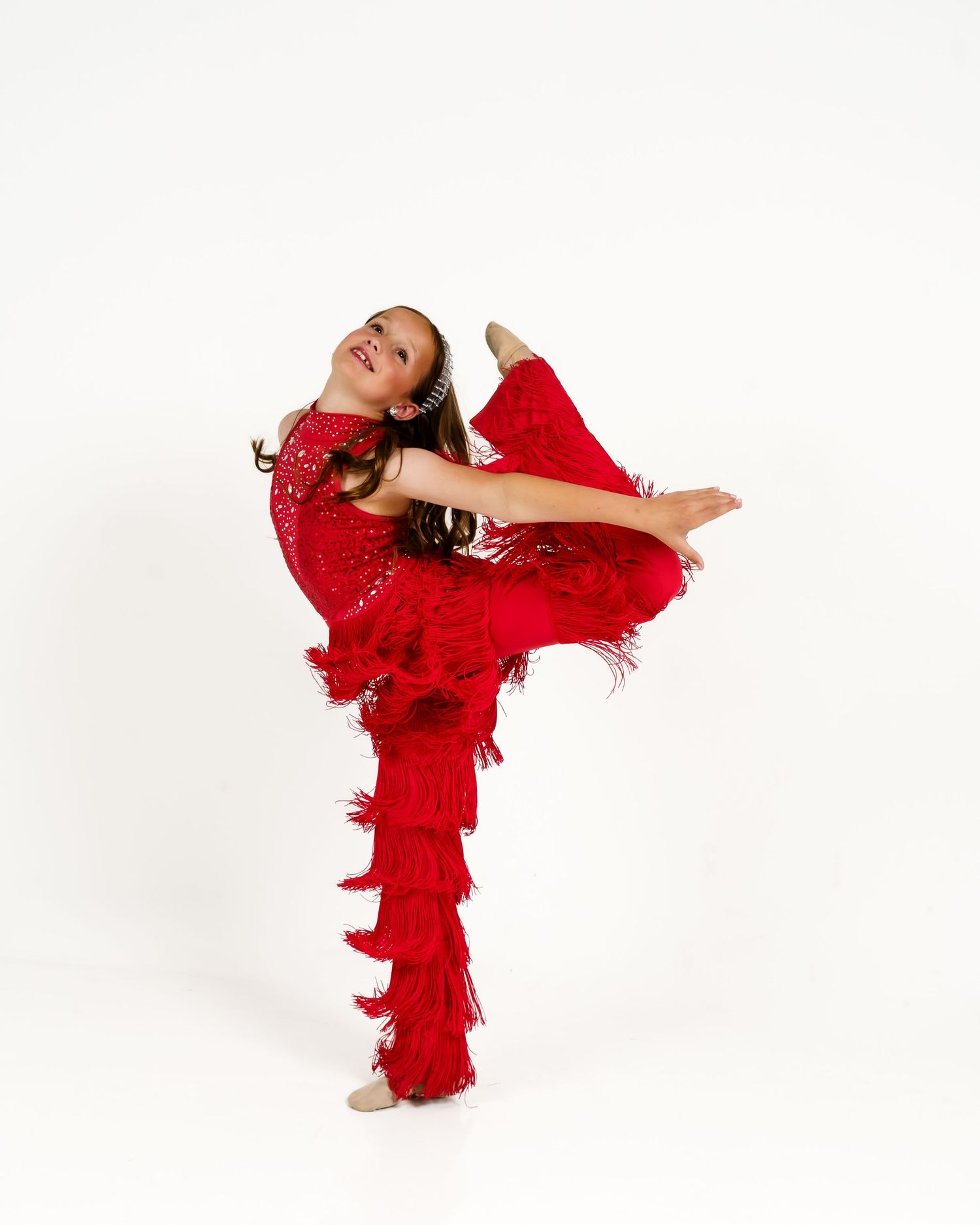 A young girl in a red fringed dance costume performs a high kick pose against a white background.
