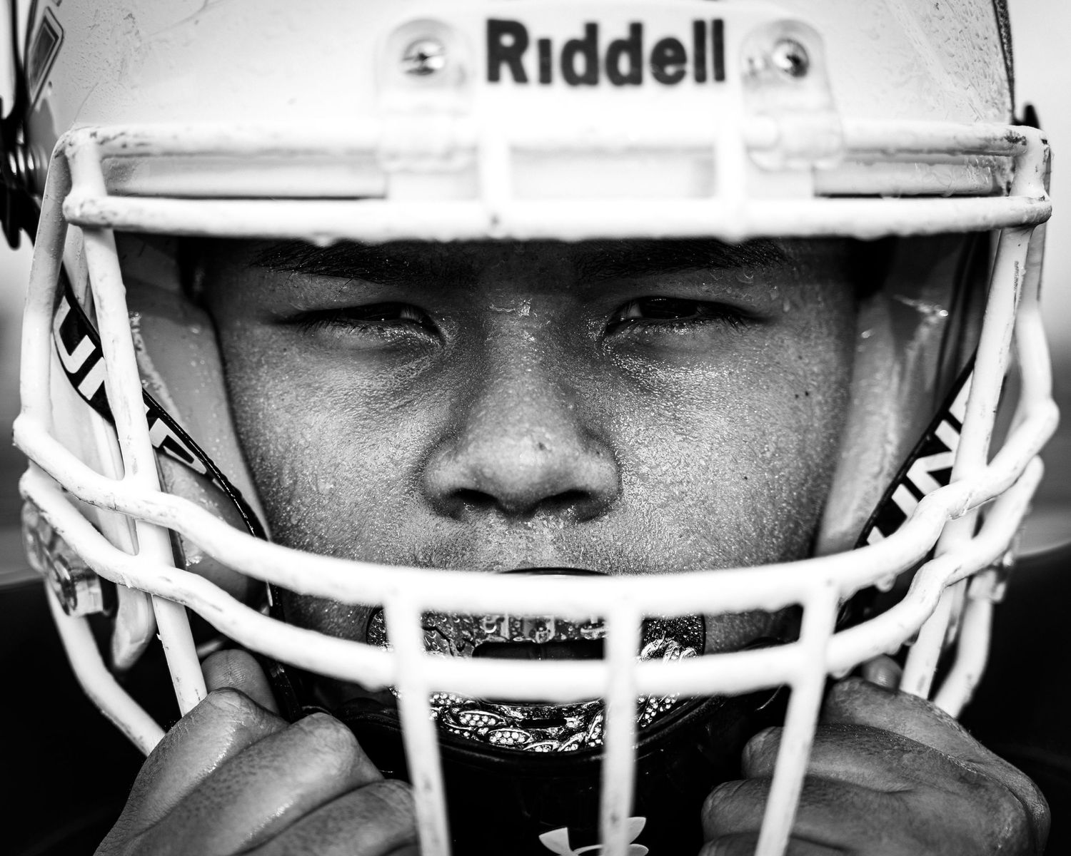 Black and white close-up of a young football player wearing a Riddell helmet, intense gaze visible through the face guard.