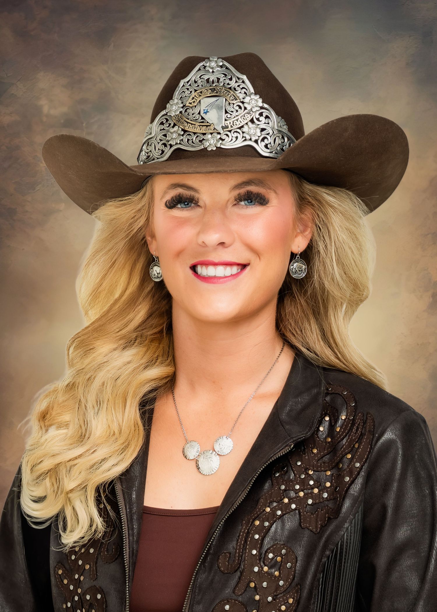 Woman in brown cowboy hat with silver band, leather jacket, and silver jewelry, smiling against studio backdrop.