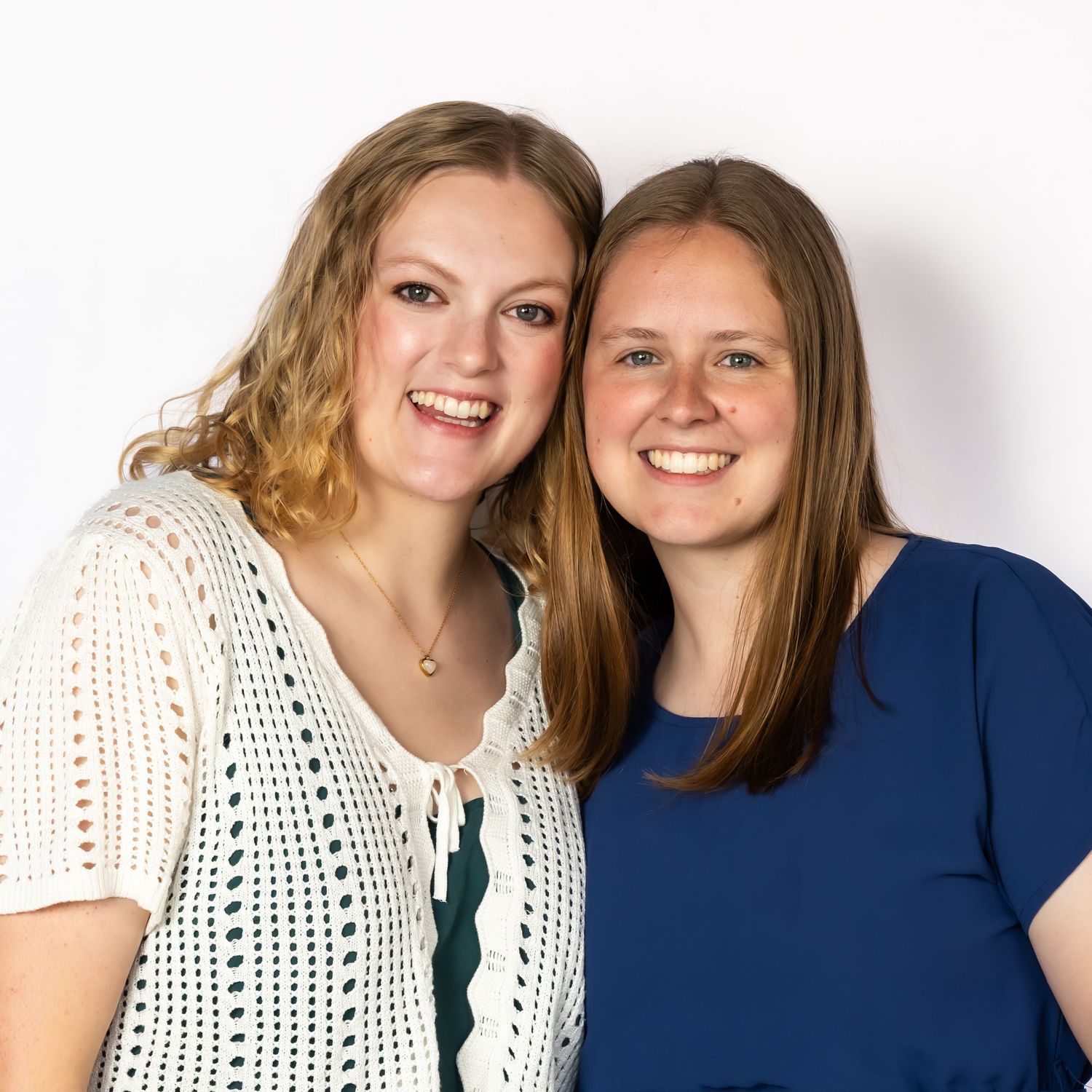 Two smiling young women posing together, one in a white lace top and one in a blue dress, against a white background.