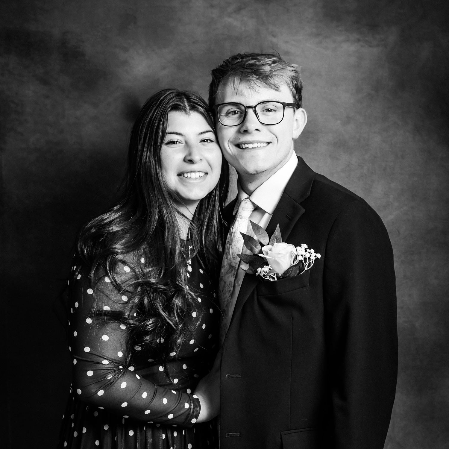 Black and white prom photo of a smiling young couple, girl in polka dot dress, boy in suit with boutonniere.