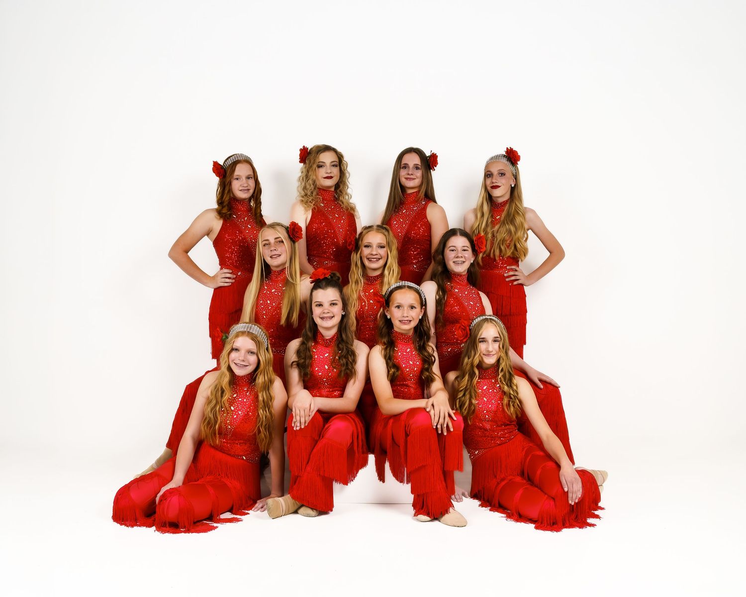 Group of young dancers in matching red dresses and floral headpieces posing together for a dance recital photo.