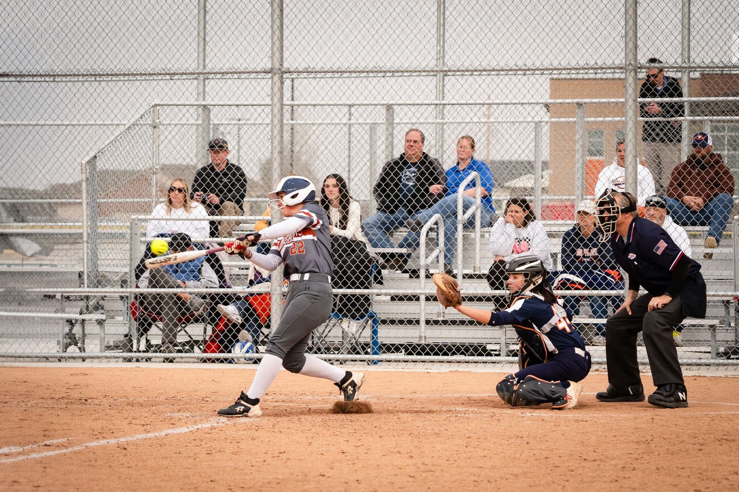 Softball batter swinging at pitch with catcher and umpire behind home plate, spectators watching from bleachers.