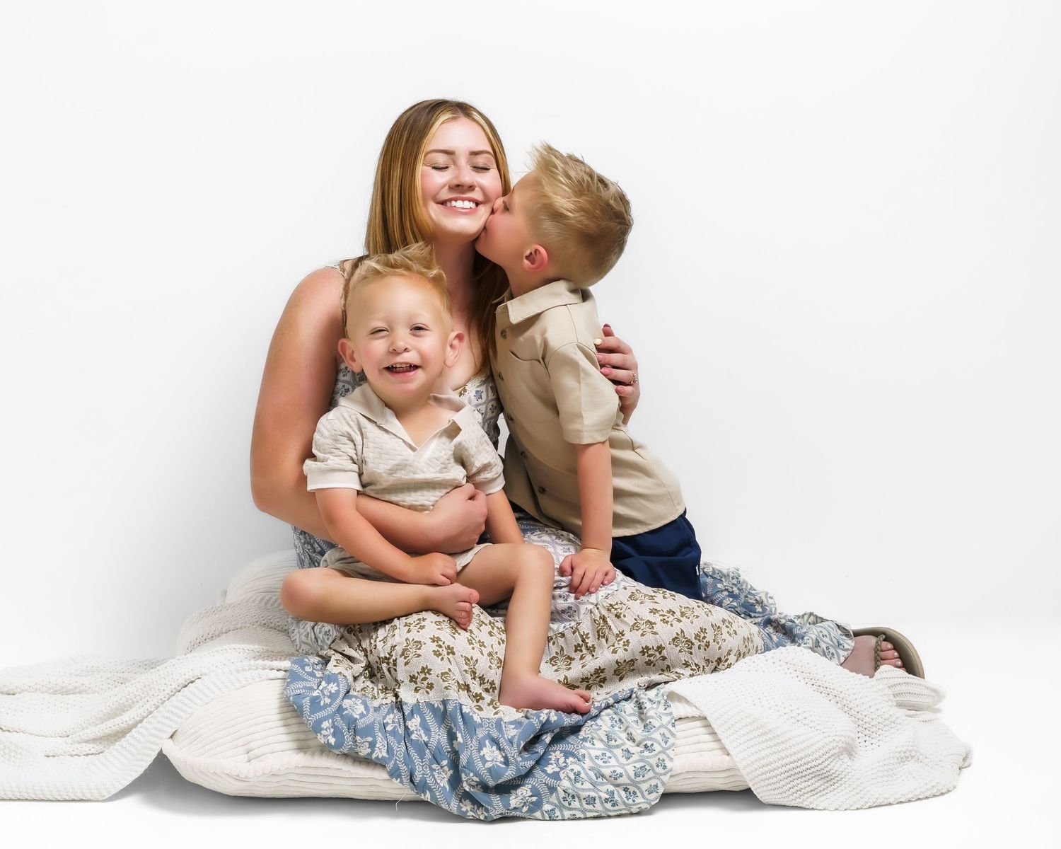 Smiling brunette mother with eyes closed as young son kisses her cheek holding toddler in studio.