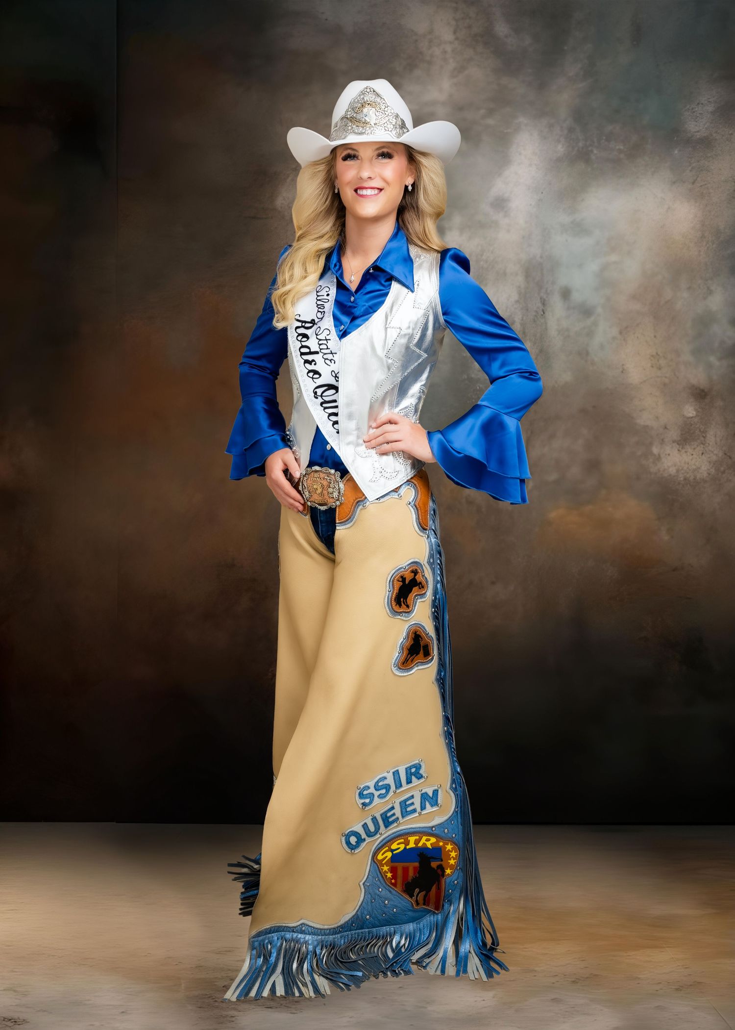 Woman in rodeo queen attire wearing white cowboy hat, blue jacket, fringed skirt, and sash at a portrait session.