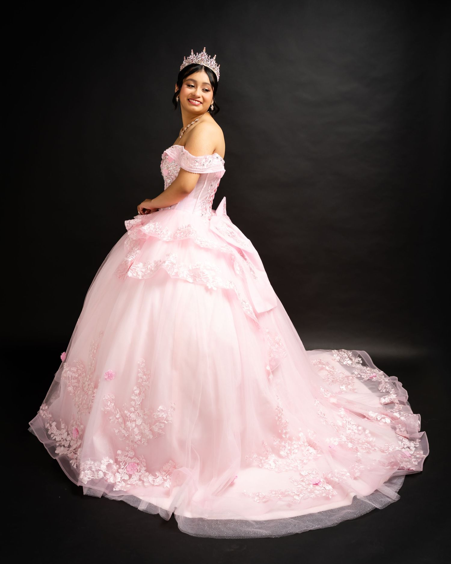Young woman in a pink off-shoulder ball gown with floral lace trim and tiara, posing elegantly against dark background.