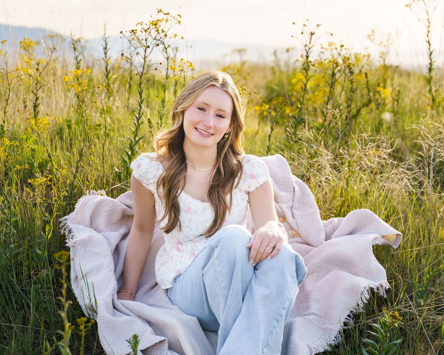 Young woman smiling in a wildflower meadow, wearing white top and light jeans with a pink blanket.