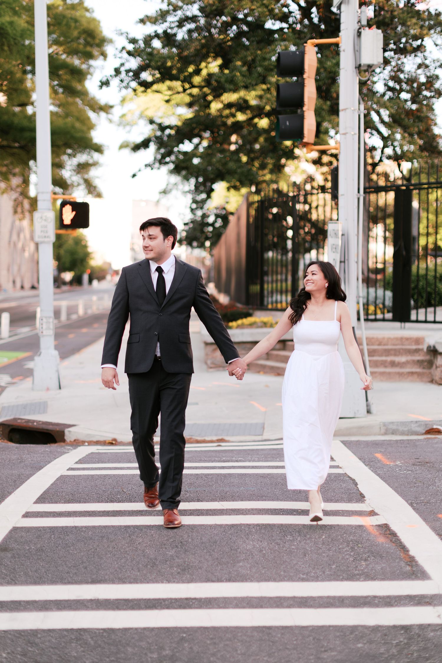 Couple in wedding attire walking hand in hand across a striped crosswalk at sunset.