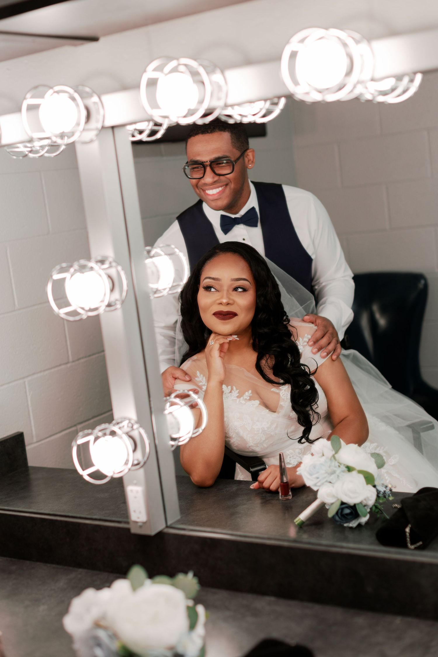Playful bride and groom pose in front of a makeup mirror with bright lights.