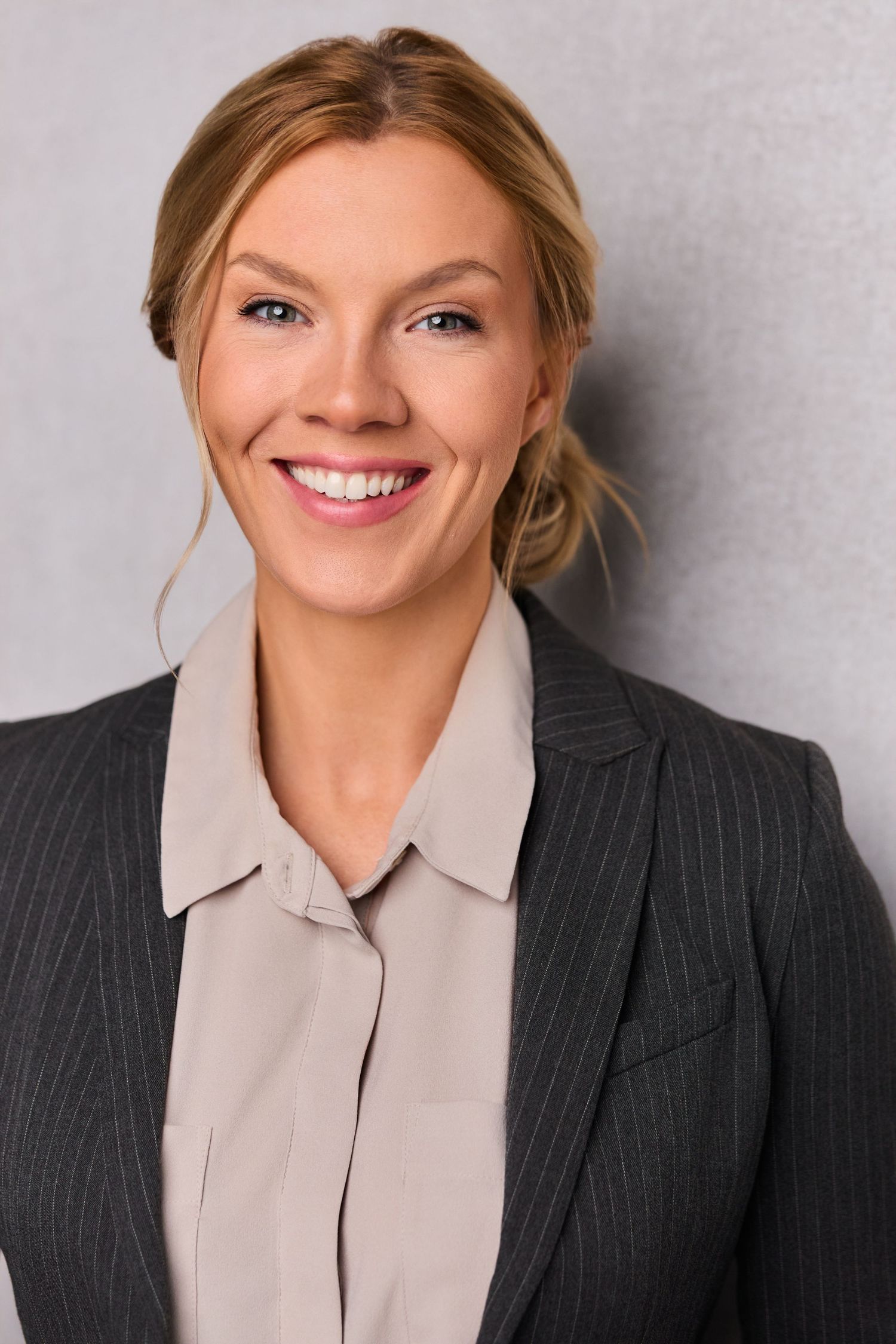 Smiling blonde businesswoman in pinstripe blazer and beige blouse poses confidently against gray background.